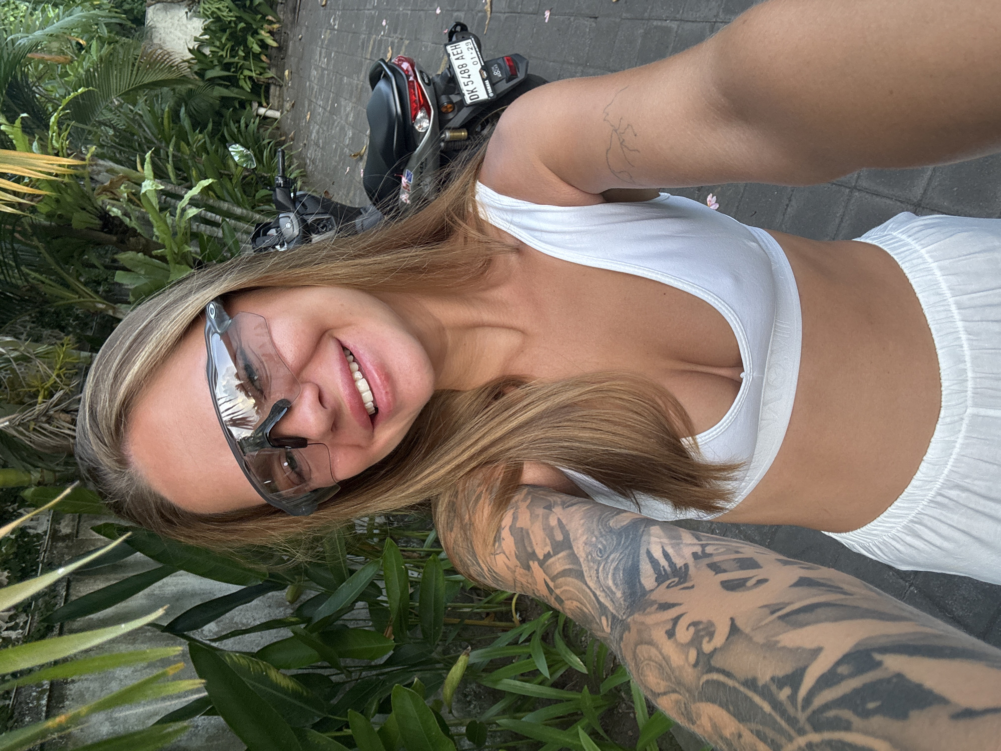 A young woman stands beside a black moped on a paved road, likely in North Kuta, Indonesia, as indicated by the vehicle's license plate "DK 5488 AEH" and sticker reading "OTMAN," commonly found on vehicles in the region. The woman is dressed in a white top and matching shorts, with clear sunglasses perched on her head. Her right arm is heavily tattooed, and a smaller tattoo is visible on her left arm. Lush tropical foliage surrounds her, suggesting a residential or tourist area. The moped is parked on the paved surface. The woman is smiling and looking directly at the camera.