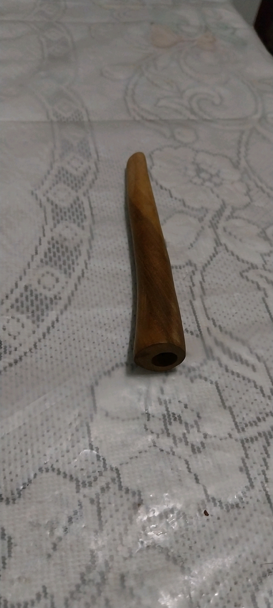 A wooden object, possibly a pipe or a mouthpiece, rests on a white lace tablecloth. The object is cylindrical with a wider end that has a visible hollow opening, and tapers to a rounded, solid end. The wood has a light brown, natural grain. The tablecloth exhibits a repeating floral and geometric pattern. The scene is illuminated by an overhead light source, casting soft shadows. The image was taken in Bojong Nangka, Indonesia, although no specific identifying landmarks are visible within the frame.