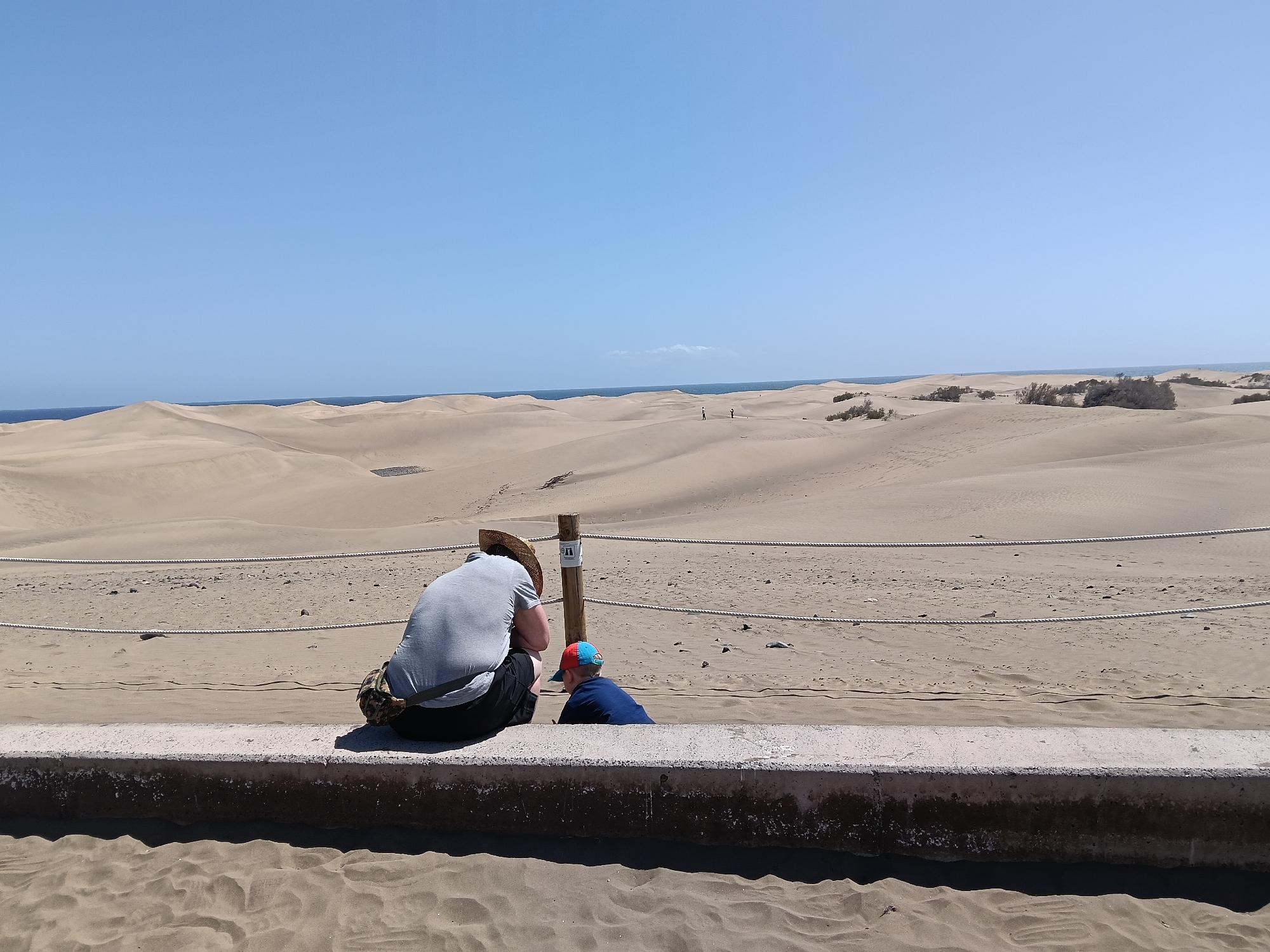 The photograph depicts a desert landscape under a clear blue sky, located in San Bartolomé de Tirajana, Spain. In the foreground, a person wearing a straw hat and a gray shirt is sitting on a concrete wall, next to a younger individual wearing a blue shirt and a red and blue cap. They appear to be observing the desert scenery, which extends to the horizon where a thin strip of ocean is visible. Ropes are strung across wooden posts behind the wall, likely to prevent people from crossing into the dunes. The ground in front of the wall is covered in sand.
