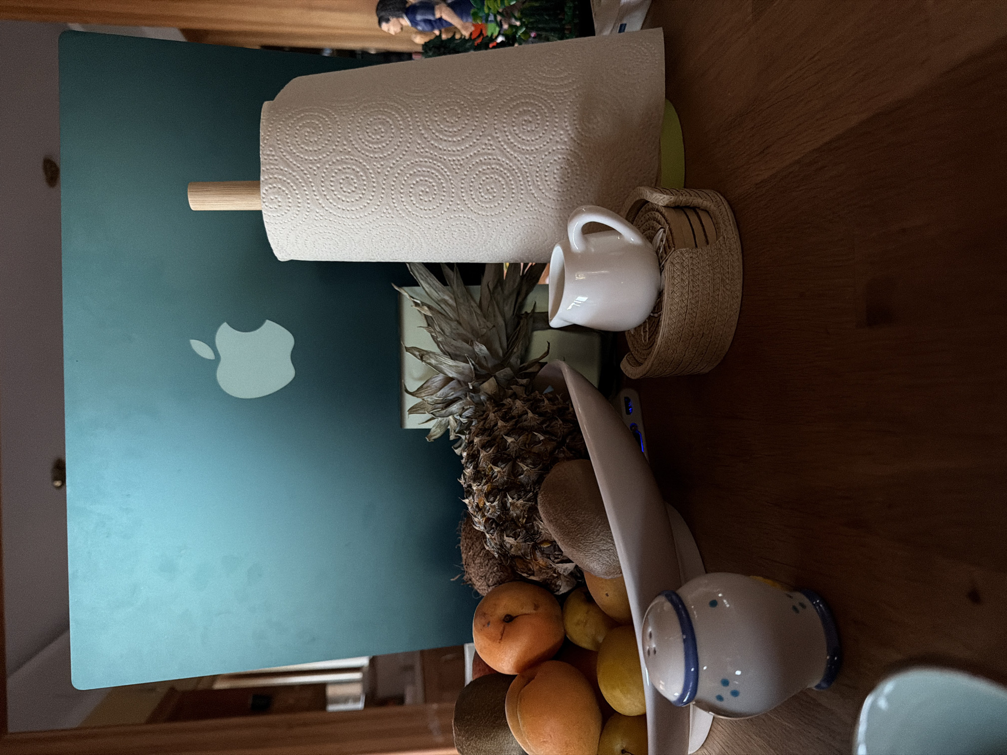 The image depicts a still life arrangement on a wooden table in what appears to be a kitchen or dining area. Dominating the background is the teal lid of an Apple laptop, with its logo visible. In front of the laptop, a roll of paper towels stands vertically, held by a wooden dowel. To the left of the paper towels, a white fruit bowl contains various fruits, including apricots, kiwis, lemons, and a pineapple. A small, white ceramic pitcher sits on a woven coaster to the right of the fruit bowl. In the foreground, a salt and pepper shaker set with blue and white detailing is visible. To the far right, a figurine of a woman in a blue outfit can be seen among some greenery. The lighting suggests an indoor setting, likely during the day, with natural light contributing to the overall illumination. The scene is static, with no individuals actively engaged in any actions. The location is described as Souni-Zanatzia, Cyprus.