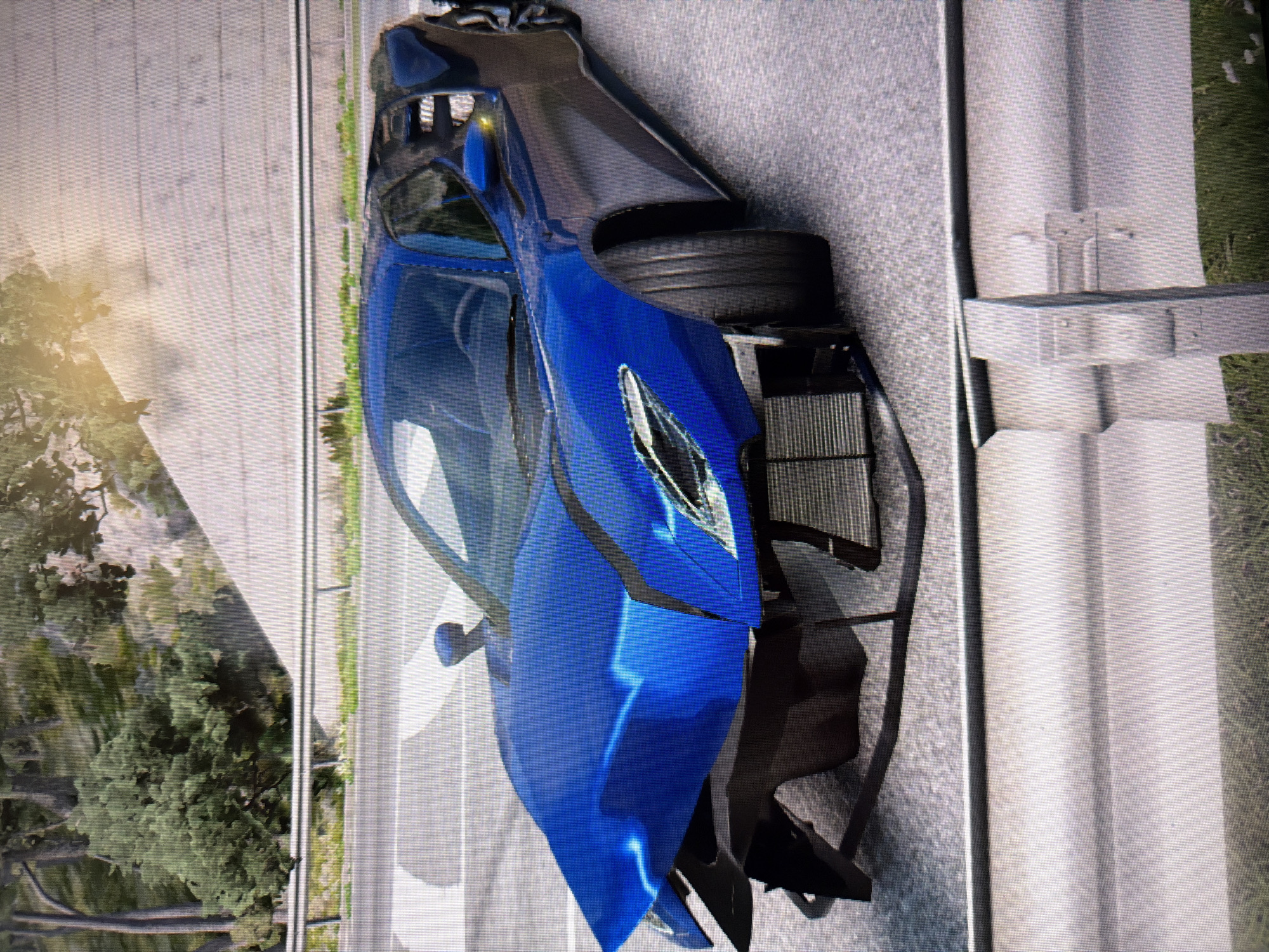 The image, captured from a screen and rotated 90 degrees clockwise, depicts a heavily damaged blue sports car. The vehicle, which resembles a modern, high-performance model, is positioned on or near a paved road with visible guardrails. The car's front end is severely mangled, with body panels missing or deformed, revealing internal components such as a radiator or intercooler assembly. One front tire is visible, detached from its wheel well and resting next to the car.
The setting appears to be outdoors during daytime, with clear weather indicated by bright lighting and a lack of precipitation. Lush green trees and foliage are visible in the background, suggesting a natural environment alongside the road. The scene portrays the aftermath of a significant car crash or severe accident, given the extent of the damage to the vehicle. There are no people visible in the image, nor is any discernible text present. The grid pattern visible across the image suggests it is a photograph of a digital display, possibly from a video game.