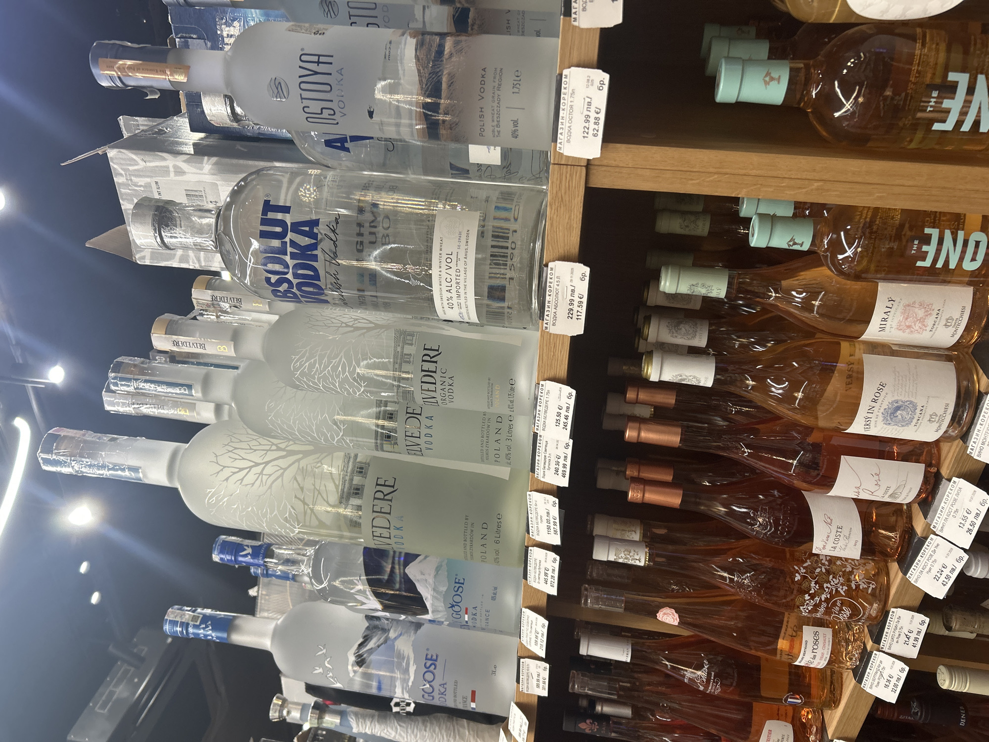 This image captures a retail display of alcoholic beverages, likely in a supermarket or liquor store in Plovdiv, Bulgaria, given the visible Bulgarian text and currency on the price tags. The shelves are stocked with numerous bottles of vodka on the left side and rosé wines on the right.
Prominently displayed vodka brands include Absolut Vodka, AL Ostoya Vodka, Belvedere Vodka (including an "Organic Vodka" variant), and Grey Goose Vodka. These are presented in various sizes, with large format bottles such as 1.75 liters, 3 liters, 4.5 liters, and 6 liters being noticeable. Product details like "POLISH VODKA," "SWEDISH WATER & WINTER WHEAT," "40% ALC/VOL," and "IMPORTED" are visible on the labels.
The right side of the image features multiple bottles of rosé wine, stacked horizontally. Brands such as MIRALÝ (Toscana), LA COSTE (Lisa, Une Nuit), VERSY IN ROSE, CHATEAU ROUBINE, and wines from BERTRAND are visible. Many bottles feature distinct labels and cap designs.
Numerous price tags are affixed to the shelf edges, displaying prices in Bulgarian Lev (лв.) and Euro (€). Each tag is labeled "МАГАЗИН-КОРЕКОМ" (MAGAZIN-KOREKOM), indicating the store's name. Examples of prices include Absolut Vodka 4.5L for 229.99 лв./117.59 €, Belvedere Vodka 6L for 1150.00 лв./587.99 €, and various rosé wines ranging from approximately 26.50 лв./13.55 € to 41.99 лв./21.41 €. Some tags also include dates, such as "11.01.2028". The bright, indoor lighting suggests a typical daytime retail environment.
