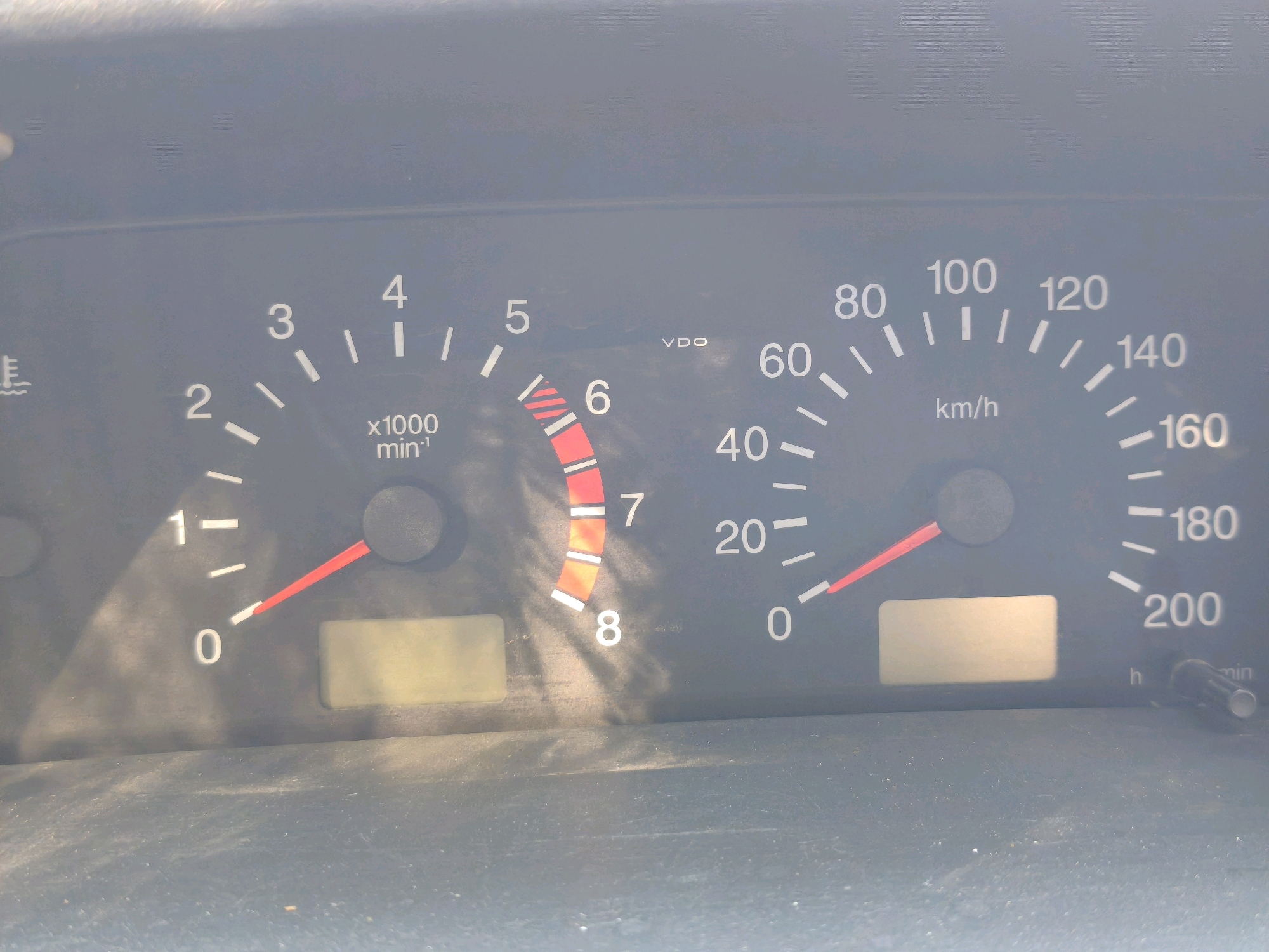The image shows a close-up of a car's dashboard, displaying the speedometer and tachometer. The speedometer, on the right, is marked in km/h and shows a reading between 0 and 200, with the needle indicating a speed of approximately 20 km/h. The tachometer, on the left, is marked in x1000 min⁻¹ and reads between 0 and 8, with the needle pointing to around 0.8. There is also a fuel gauge or a temperature gauge which is not clearly identifiable. The dashboard is dusty. The background is out of focus and appears to be the interior of a car. There is no indication of the location being Karachayevsk, Russia, or any urban environment. The image is a static capture of the dashboard.