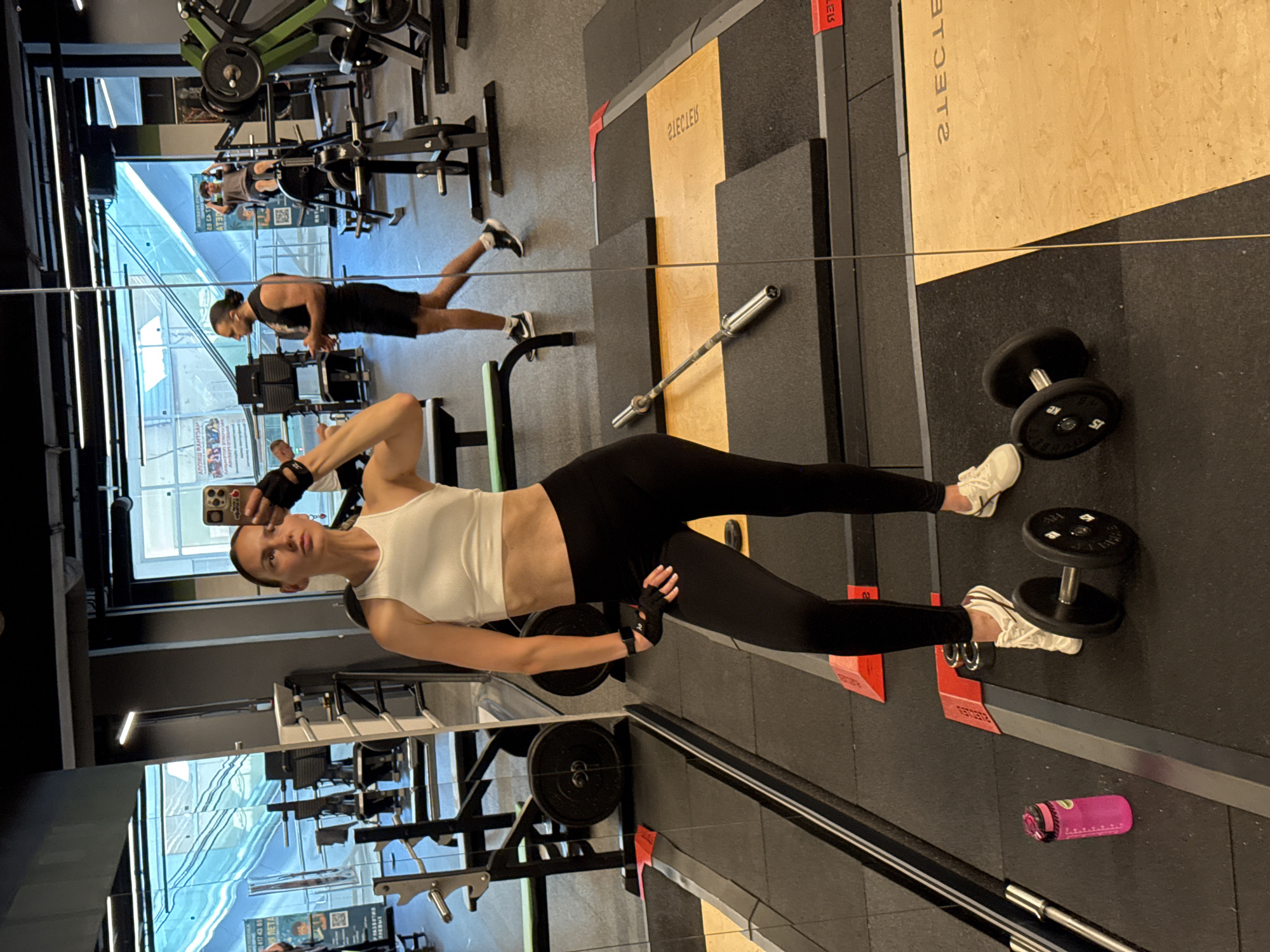 A woman is taking a mirror selfie in a gym. She is wearing a white crop top and black leggings, and she has on black weightlifting gloves. She is holding a smartphone in her right hand and has her left hand on her hip. In the reflection, a man wearing a black tank top and shorts with headphones in his ears is walking away from the camera. In the background, gym equipment is visible, including weight machines and barbells. The gym has large windows that look out onto an escalator or ramp. The floor is covered in black rubber mats, with some wooden platforms. The overall impression is a modern and well-equipped fitness facility.