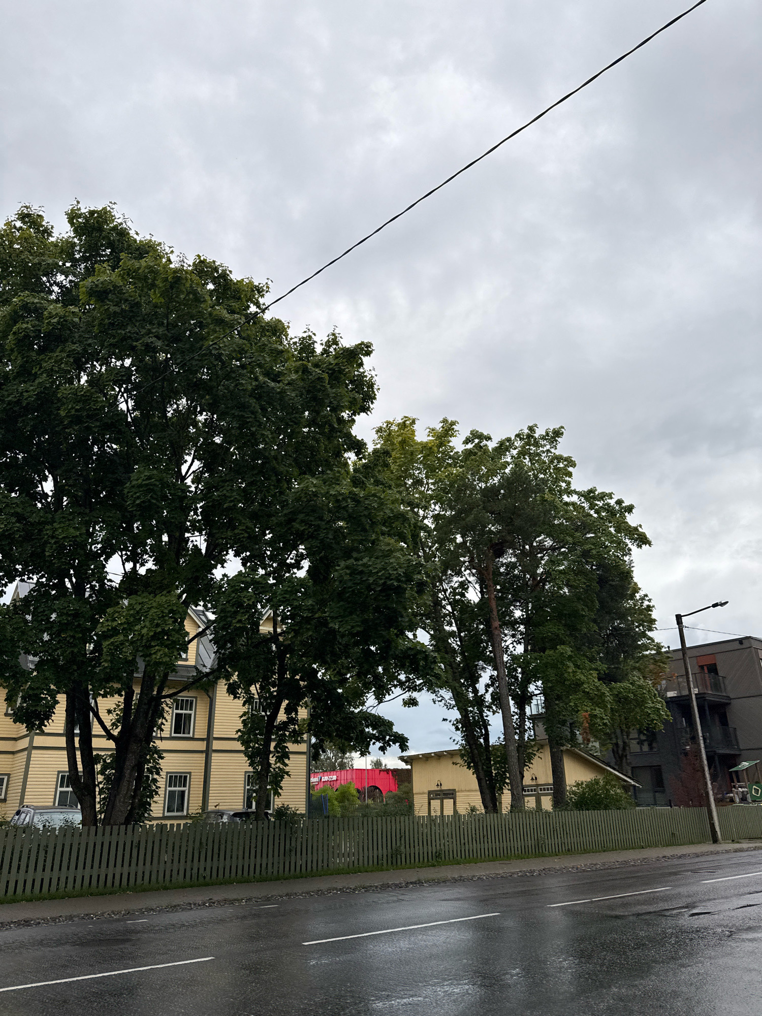 The image depicts a street scene in Tallinn, Estonia. On the left side of the frame, a weathered, pale yellow wooden house with white-trimmed windows is visible, partially obscured by lush green trees. A dark green wooden fence runs along the foreground, separating the sidewalk from the property. Beyond the yellow house, a modern, dark-colored apartment building with balconies stands on the right, topped by a streetlamp. The road in front is wet and reflects the overcast sky, suggesting recent rain. The sky itself is a uniform grey, indicating cloudy weather.