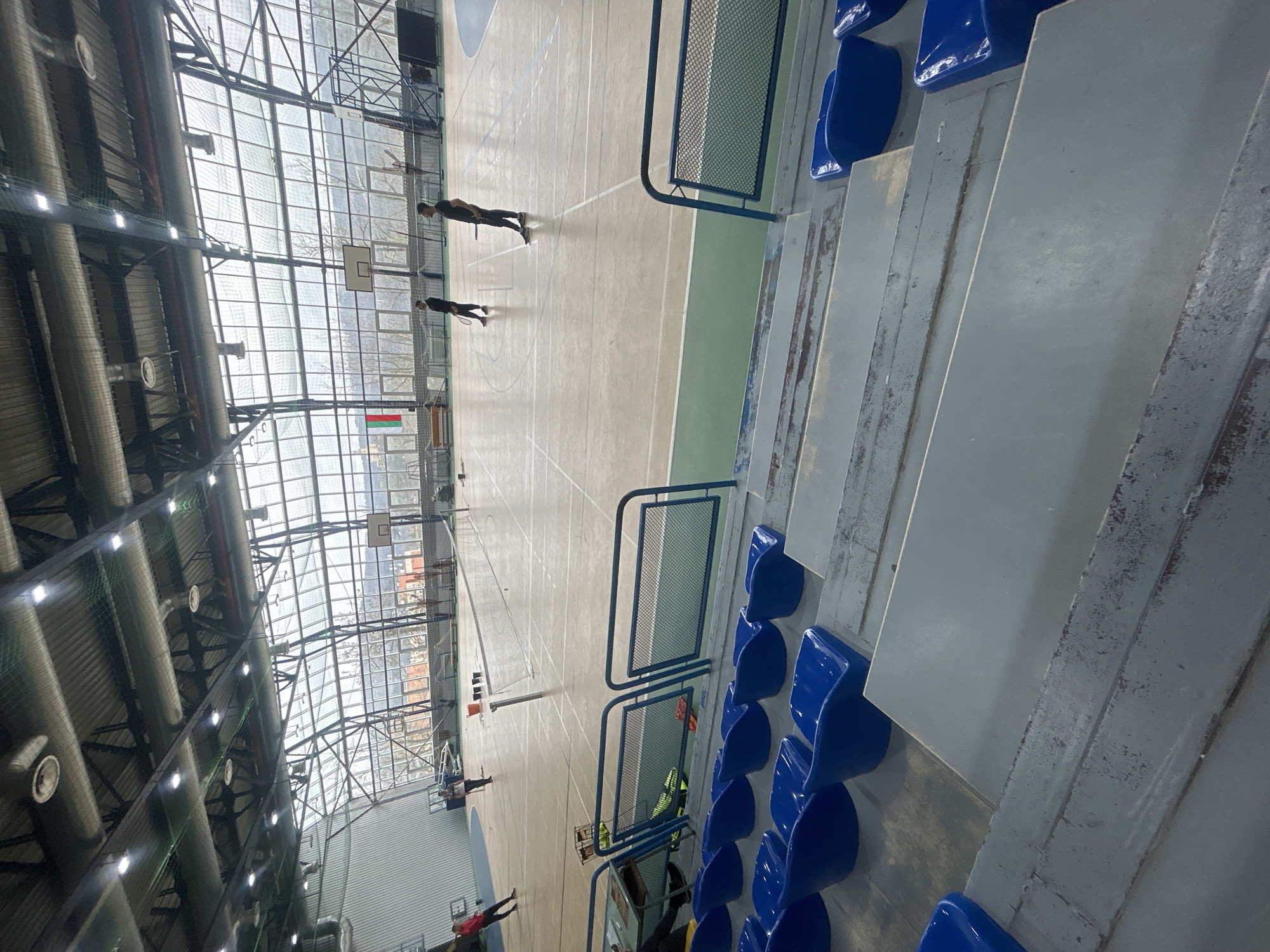 The image depicts an indoor sports hall, likely located in Veliko Tarnovo, Bulgaria. The hall has a high ceiling with exposed structural elements and translucent panels allowing natural light to enter. The floor is marked with lines indicating a basketball court, and a badminton net is set up across the court's width. Several individuals are present, appearing to be engaged in badminton. Seating is arranged along the side, consisting of rows of blue plastic chairs. The overall environment appears functional and well-lit.