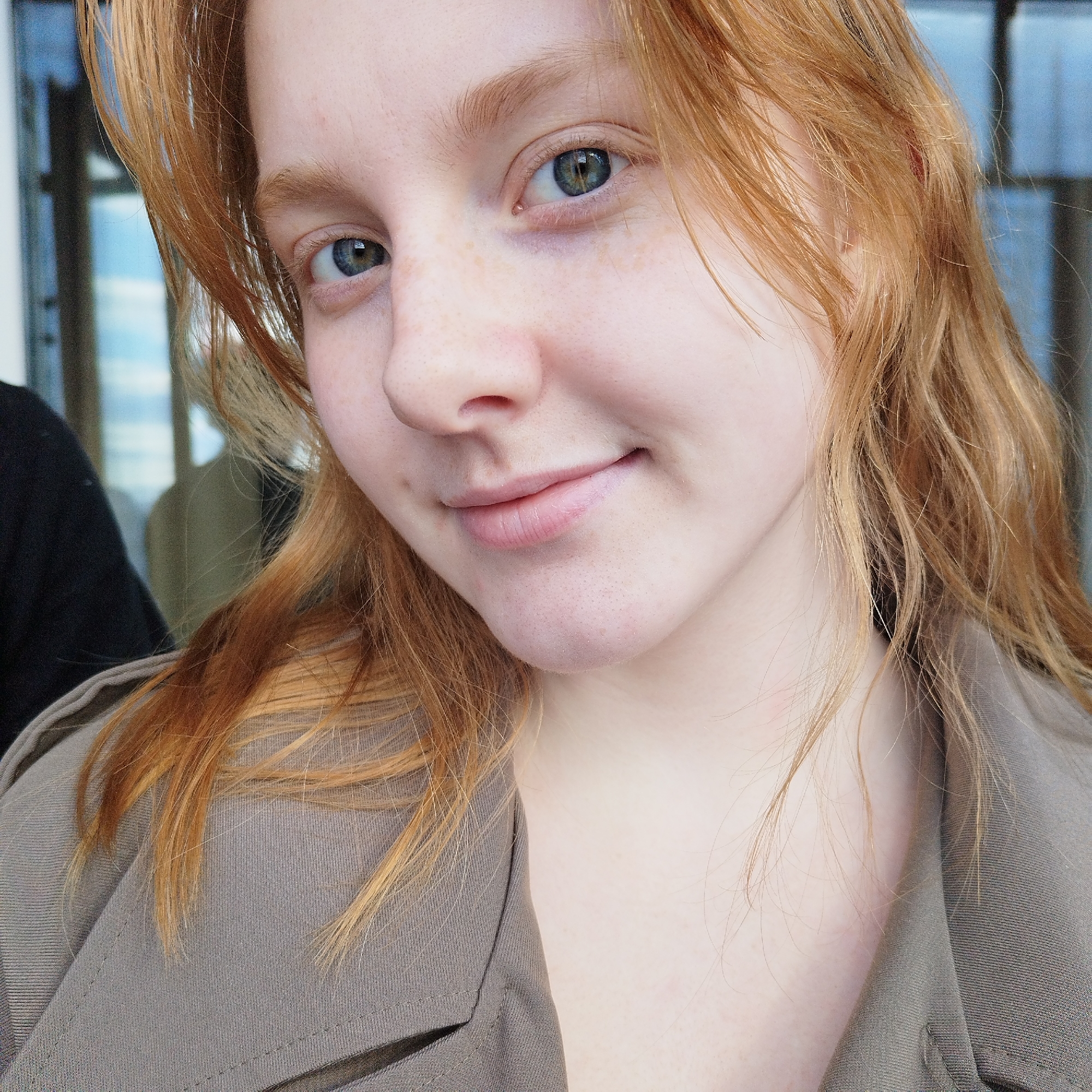 A close-up, eye-level portrait captures a young woman with fair skin and reddish-blonde hair. The woman is seen from the chest up, wearing a taupe-colored collared jacket. Her eyes are light blue and she has freckles across her nose. She has a slight, closed-lip smile. Part of another person wearing a black shirt is visible on the left side of the frame. The background shows an out-of-focus building with blue-tinted windows, likely suggesting an indoor location near a window.
It is not possible to provide a precise location in Yekaterinburg, Russia from this image alone. More information about the setting would be required to establish a location.