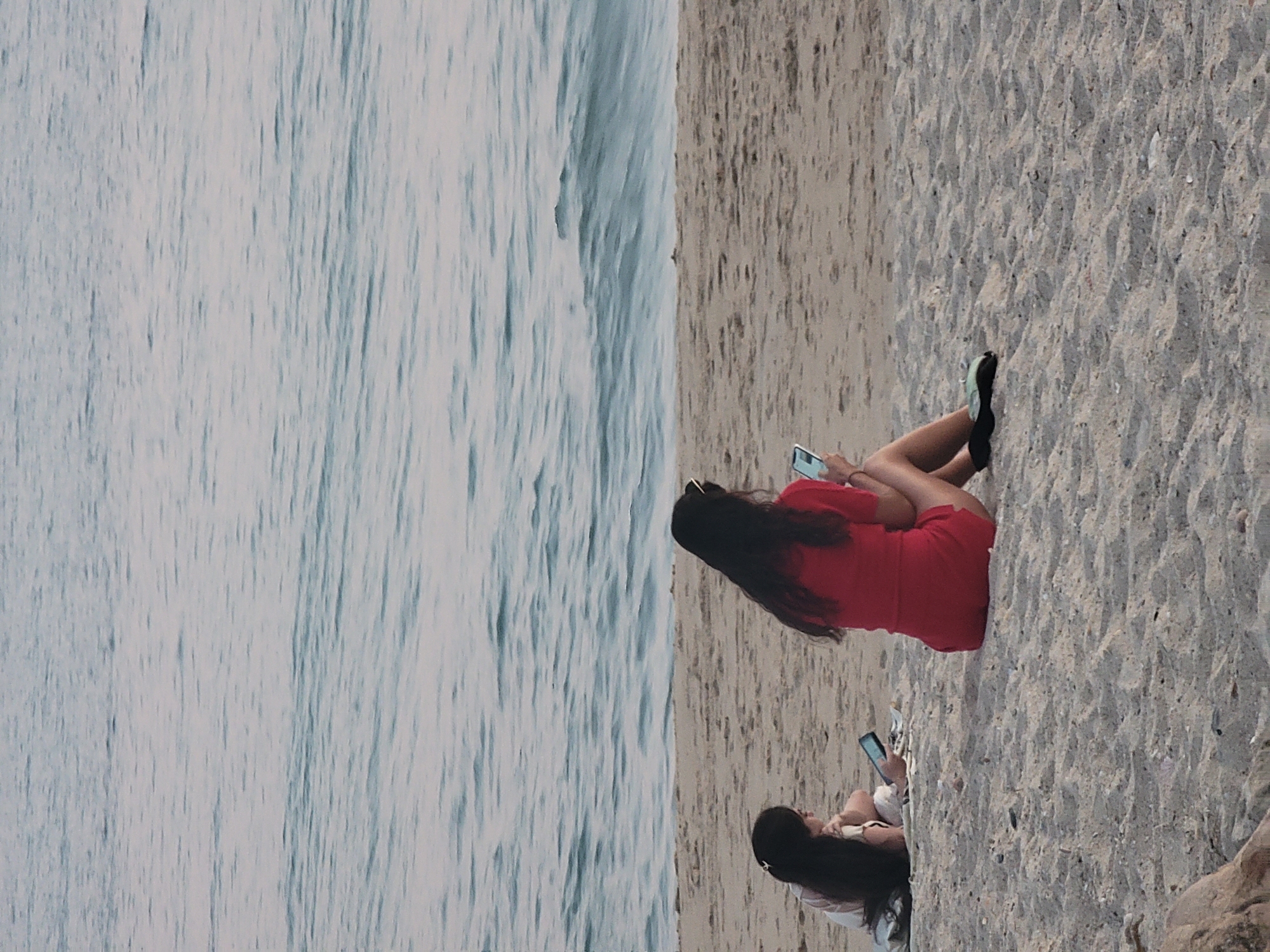 Two women are sitting on a sandy beach, looking at their phones. The woman on the right is wearing a red dress and the woman on the left is wearing a light-colored top and shorts. They are both facing away from the camera, towards the ocean. The ocean is calm with small waves. The sand is light brown and there are a few small rocks scattered around. The scene appears to be in Quarteira, Portugal, known for its beaches.