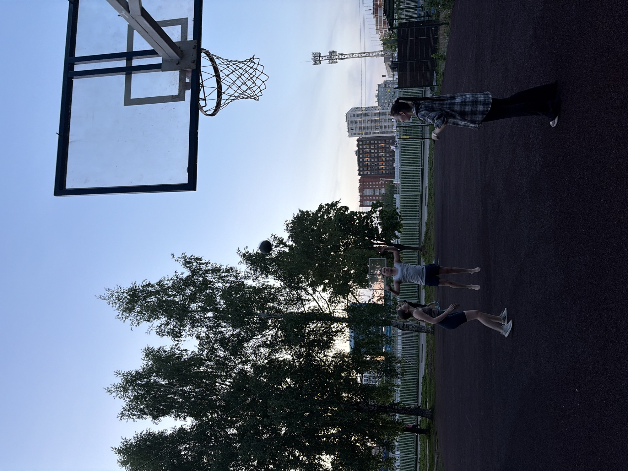 The image depicts a basketball court in Izhevsk, Russia, with a hoop in the upper left. Two people are playing basketball; one person is shooting and another is running. A woman wearing a long-sleeve shirt and dark pants is standing at the edge of the court. The background includes buildings, fences, and trees. The sky is clear. A sports complex tower can be seen in the distance.