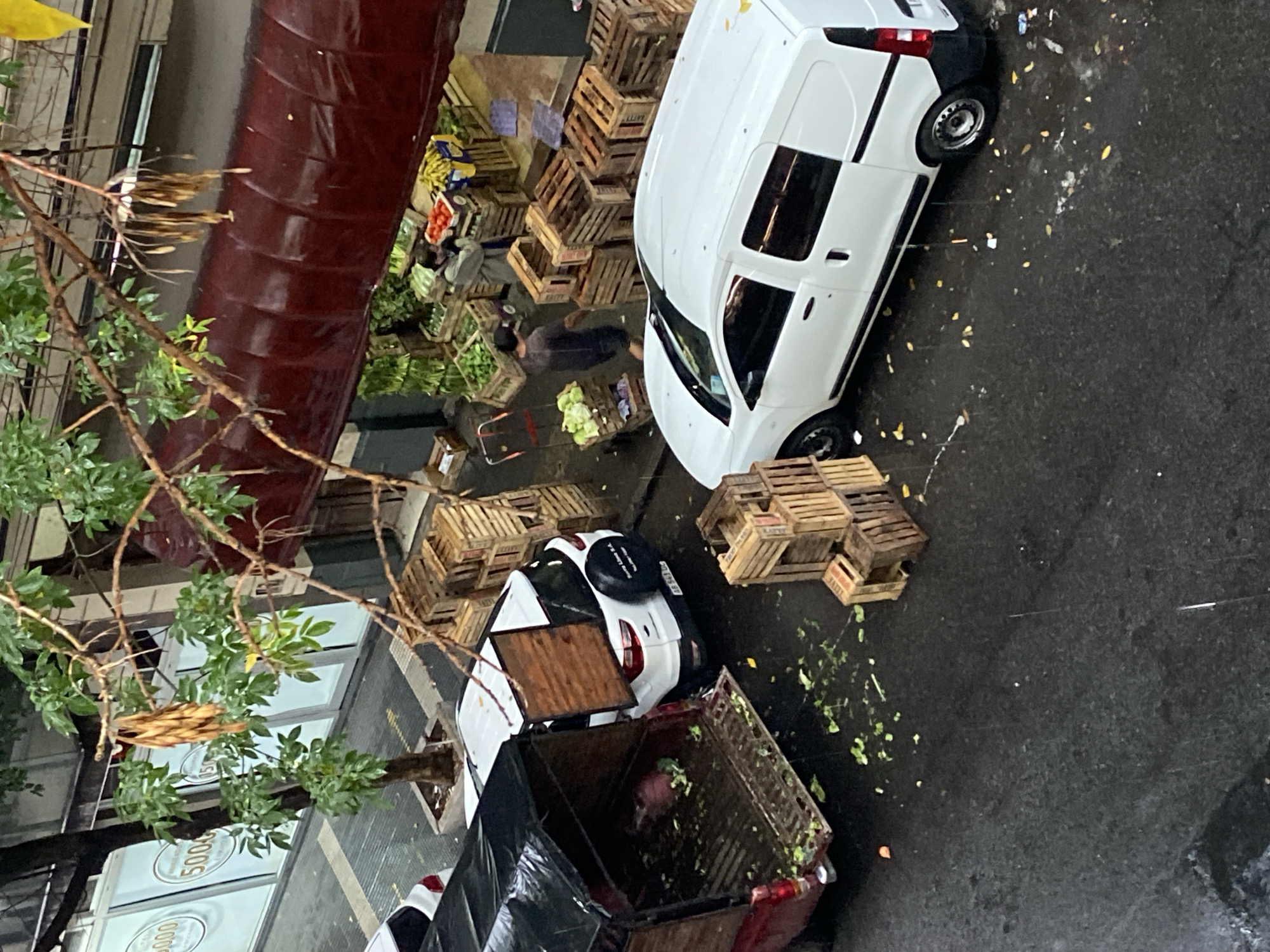An elevated, angled view shows a Buenos Aires street scene dominated by commerce. A white van and a white car are parked on the right. Numerous wooden crates, some stacked, suggesting a market or delivery point, are scattered around the vehicles. Some crates are labeled "KALTY."
Two indistinct human figures are near the crates on the left. A red truck with a black tarpaulin covering its contents is on the bottom left of the frame. Green leafy debris is scattered on the wet pavement. A tree with sparse foliage is present. A building with a dark red facade runs along the left side of the frame. Signs bearing the number "5000" are visible on the side of a building. The environment appears wet.