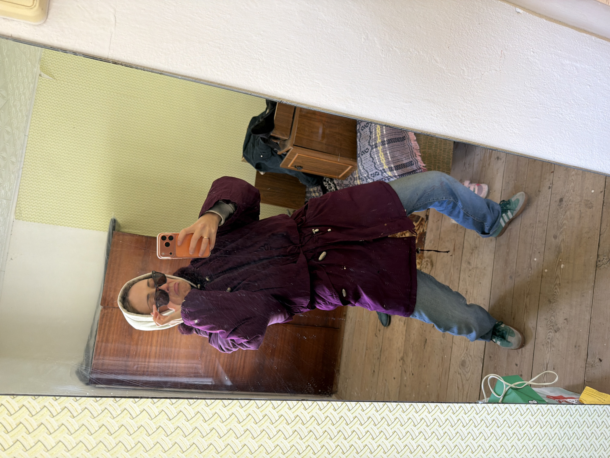 A person stands reflected in a mirror, taking a selfie with a smartphone. The person is wearing sunglasses, a hooded sweatshirt, a maroon coat, and jeans, with green sneakers on their feet. The mirror's reflection reveals a room with patterned wallpaper and wooden floors. Furniture, including a dresser and small cabinet, is visible. In the background, there is a brown patterned blanket. At the bottom of the mirror, there is also patterned wallpaper. In the bottom-right of the mirror's reflection, there is a green package with a white string and a yellow piece of paper, sitting on the wooden floor. The room appears to be old and perhaps in need of repair. Kamyshin, Russia cannot be confirmed from the image.