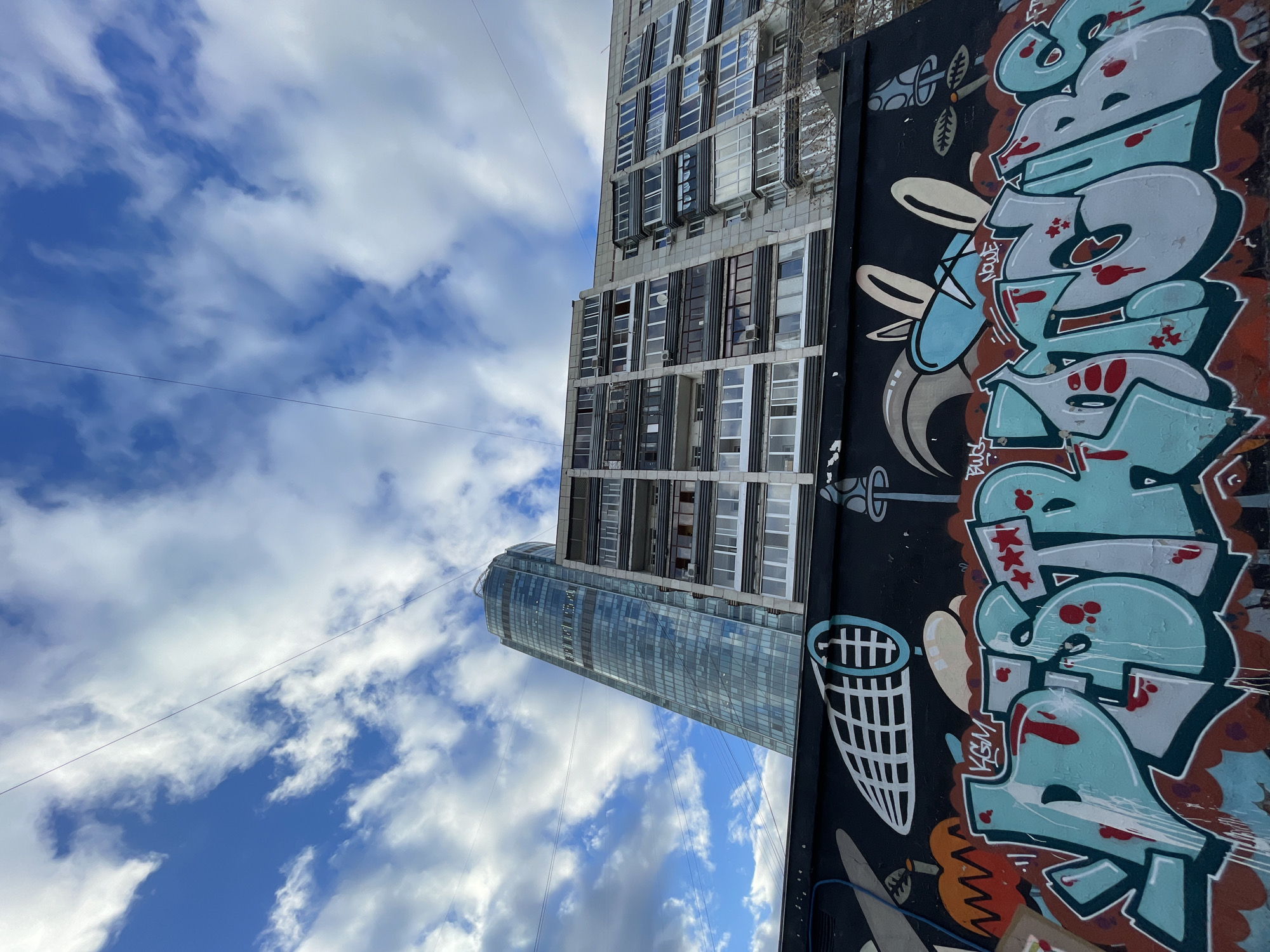 The image presents a view upwards towards a cloudy sky, capturing elements of urban architecture and street art. A building with a glass facade stands alongside a multi-story building with rows of windows. Adjacent to these buildings is a wall adorned with colorful graffiti.
The location is Yekaterinburg, Russia.