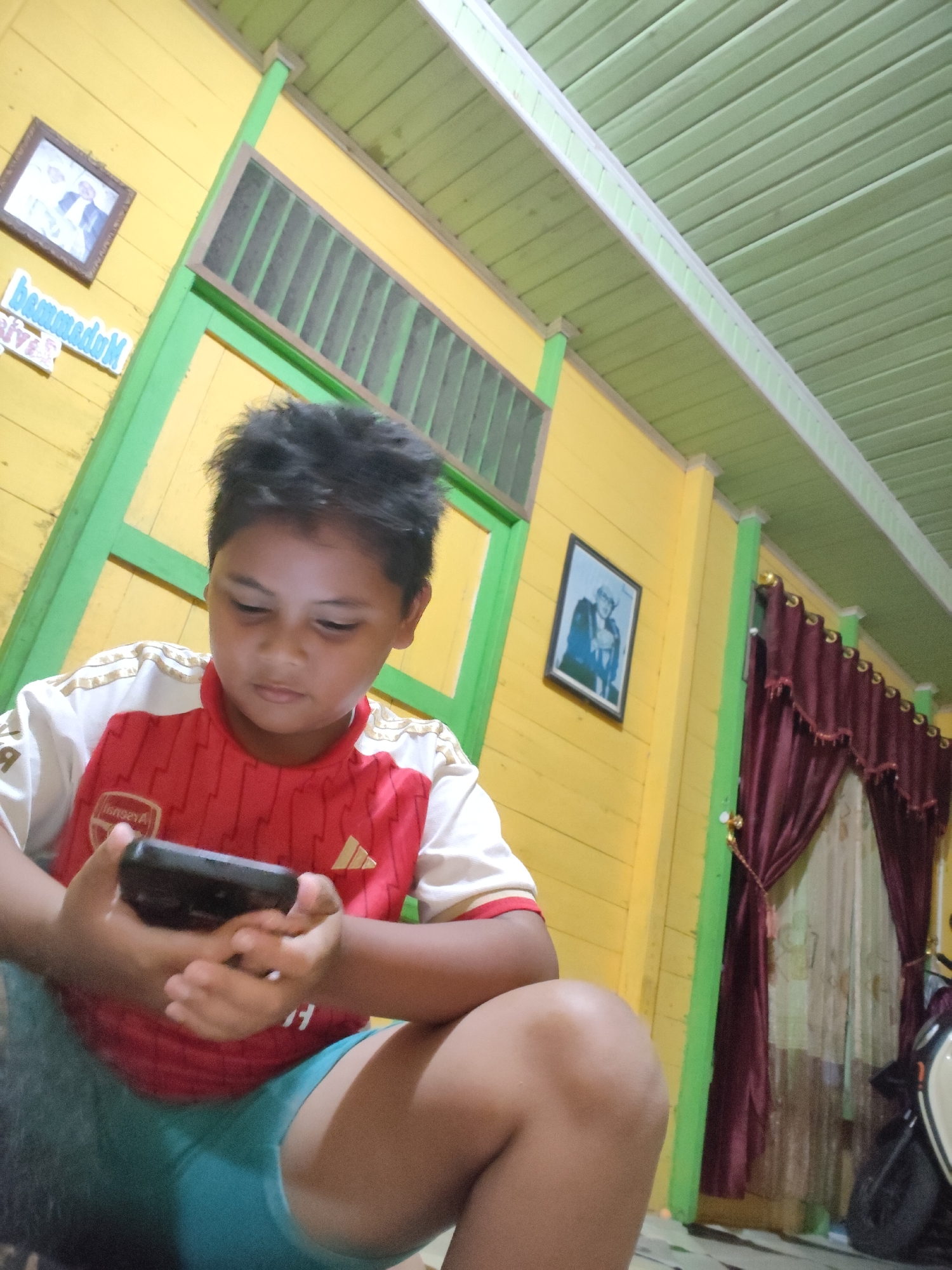 A young boy wearing a red and white shirt and blue shorts is sitting and looking at his mobile phone. He is indoors, in a room with yellow walls and a green ceiling. There are framed pictures on the wall, a green window with slats, and maroon curtains hanging near a window. The image appears to have been taken in Tanah Grogot, Indonesia.