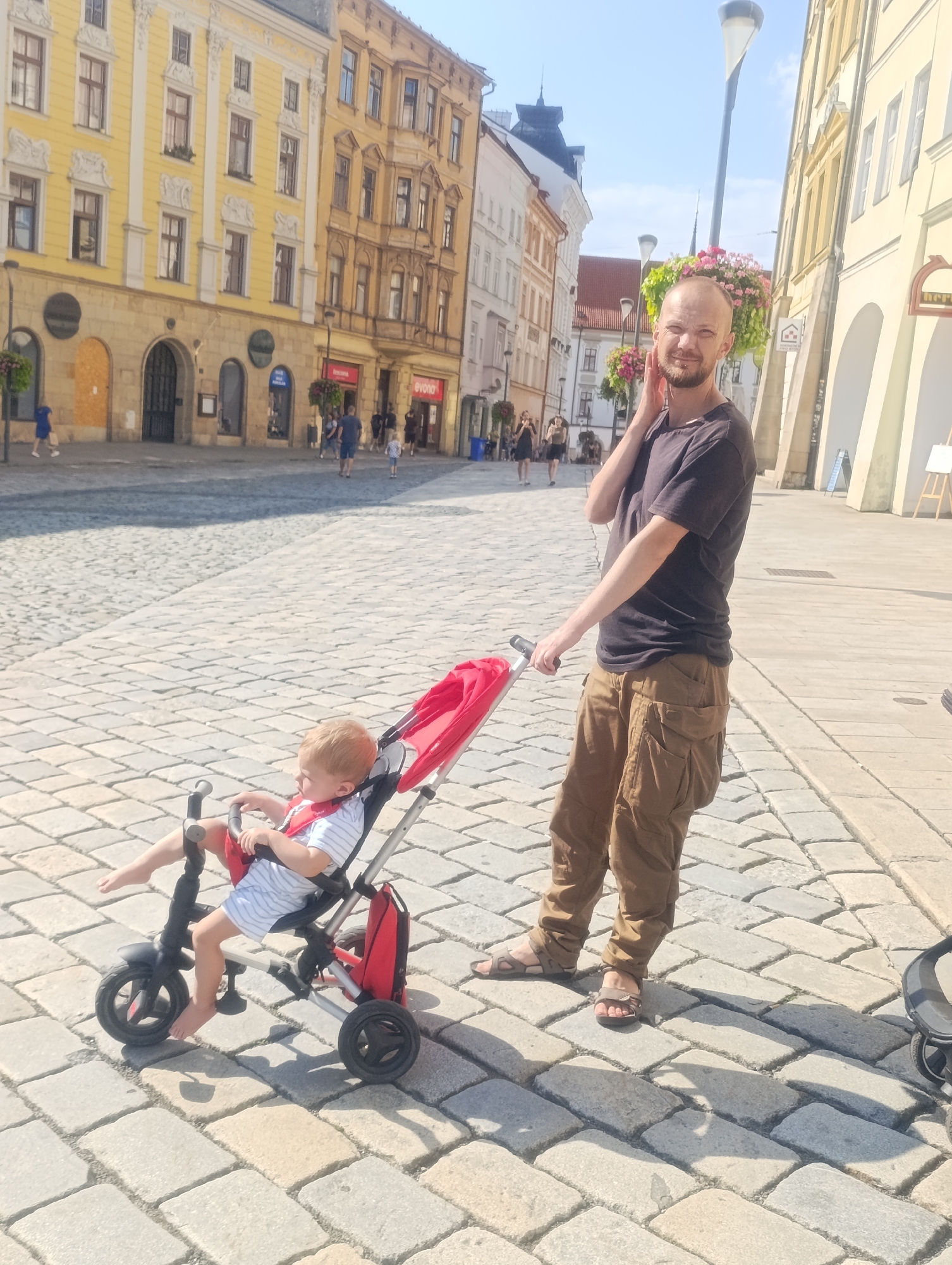 In the city of Olomouc, Czechia, a man stands on a cobblestone plaza, holding the handle of a red stroller. A young child, wearing a striped shirt, sits in the stroller, with one leg extended. The man, bald with a beard, is looking towards the camera and has his hand on his neck. He is wearing a dark t-shirt and brown cargo pants. The stroller is a tricycle-style stroller with three wheels, and the child is secured with a red harness. In the background, there are historic buildings with yellow facades, an outdoor lamp with flower baskets, and other pedestrians walking in the distance. The scene is bathed in bright sunlight, casting shadows on the cobblestones.