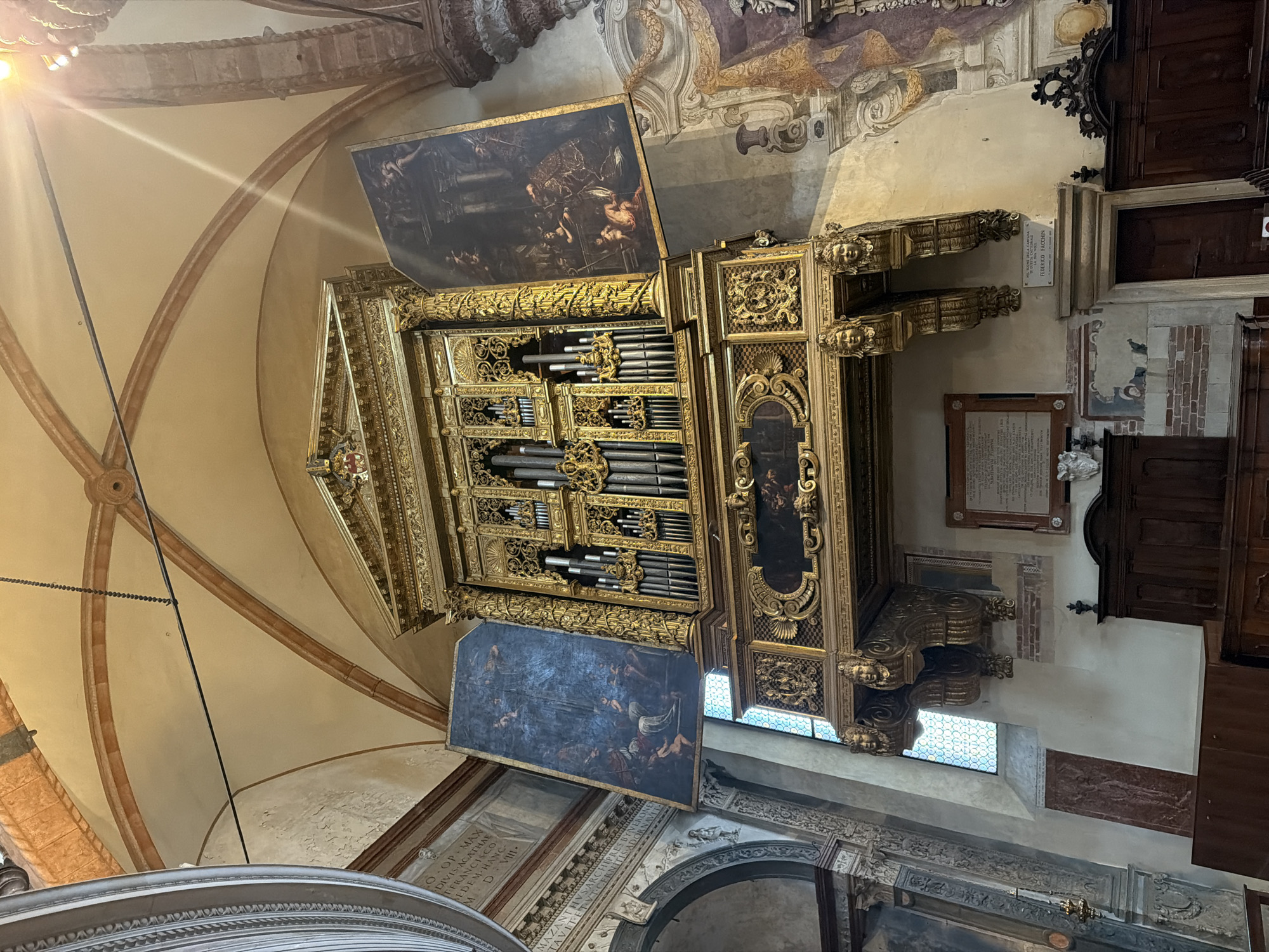The image shows a grand pipe organ located inside a church in Verona, Italy. The organ is ornately decorated with gold leaf and is flanked by two large paintings. The paintings depict religious scenes and are hung on either side of the organ's housing. The organ itself has numerous pipes of varying sizes, arranged in an organized fashion. Below the organ, there is a decorated shelf with carved details. Two carved cherub heads support the shelf, acting as corbels. A plaque with text is mounted on the wall below the shelf. In the lower part of the image, wooden pews are visible, suggesting the organ is part of a church's worship space. The ceiling of the church features arched vaults with decorative brickwork. The overall impression is one of historical grandeur and religious significance.
