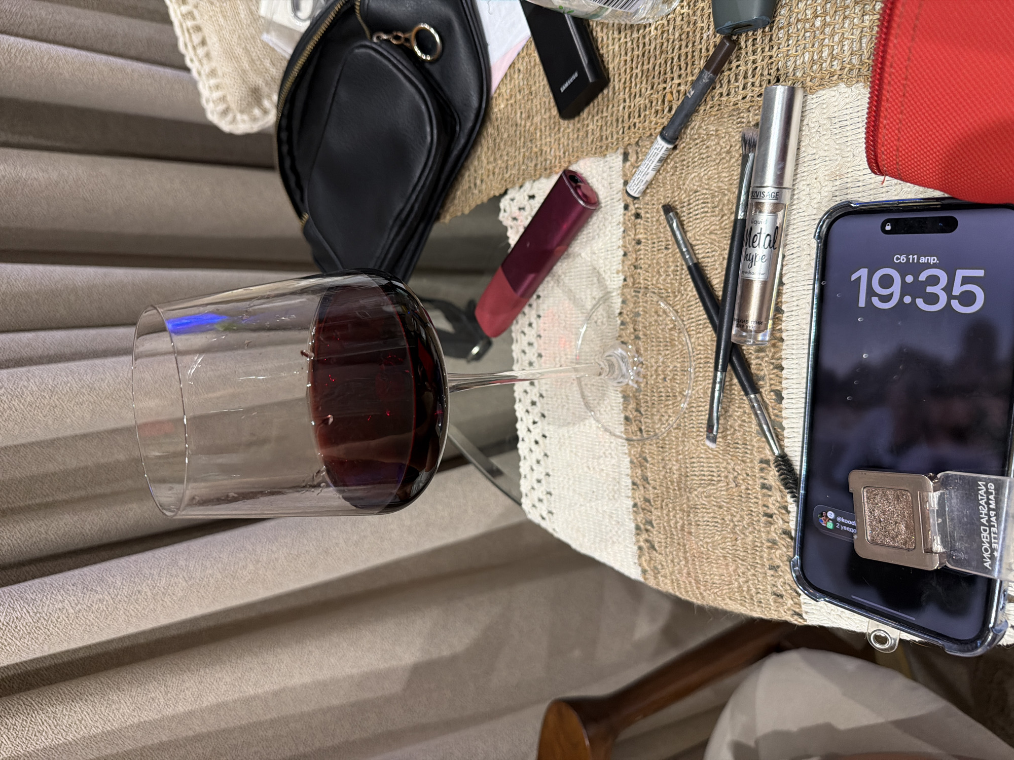 A wine glass containing red wine sits on a table alongside several cosmetic items, including brushes, eye shadow, and lipstick. A mobile phone displays the time 19:35 and text in Russian. The environment includes a textured table surface and a background with horizontal blinds. No people are visible in the image.