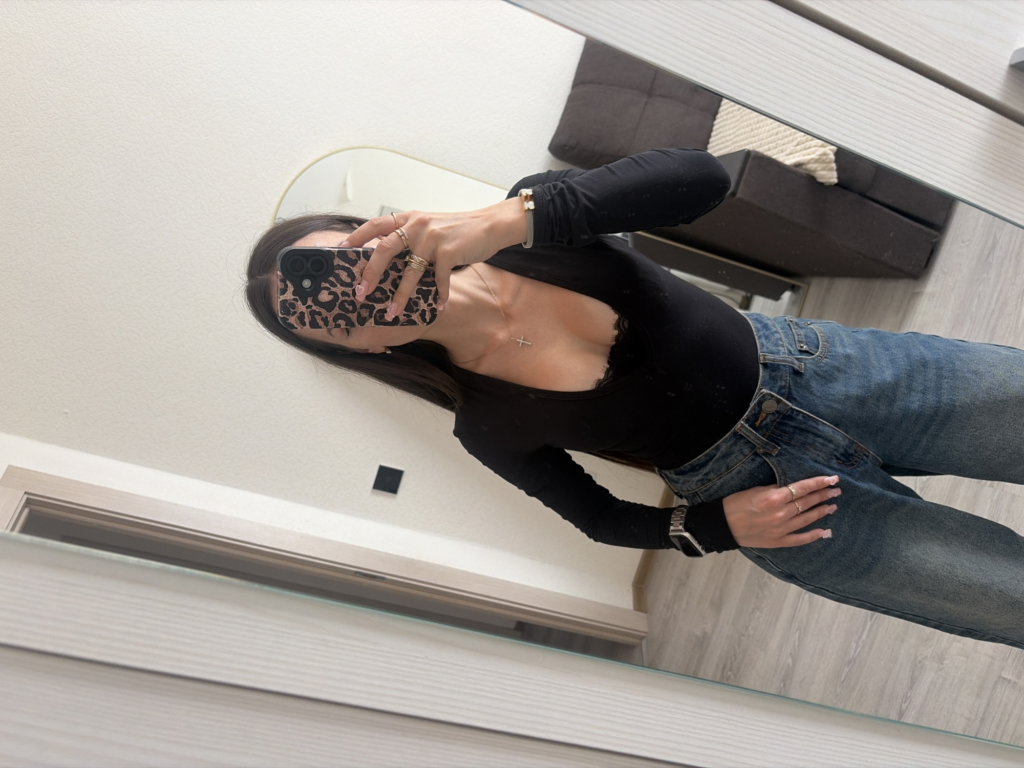 A person is taking a mirror selfie in an indoor setting. They are wearing a black long-sleeved top with lace trim, blue denim jeans, a silver smartwatch, a gold necklace with a cross pendant, a gold bracelet, and multiple gold rings. The person is holding a smartphone with a leopard-print case. In the background, a mirror reflects a doorway and a sofa. The floor appears to be light-colored wood. The overall ambiance suggests a residential interior. There is no information to place this image in Saint Petersburg, Russia.