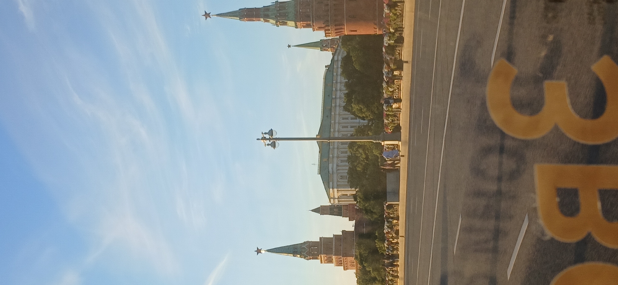 The image depicts a wide-angle view of a cityscape under a bright, pale blue sky with wispy clouds. In the background, a large, light-colored building with a green roof is visible, flanked by two prominent red towers topped with golden stars. These towers, characteristic of Kremlin architecture, are partially obscured by lush green trees. A lamppost stands in the middle ground. In front of the building and trees, a stone wall with decorative elements and several small figures can be seen. A road with white lane markings occupies the foreground, with blurred yellow lettering on a reflective surface visible at the very bottom of the frame. The scene appears to be captured from a slightly elevated perspective, possibly from a moving vehicle.