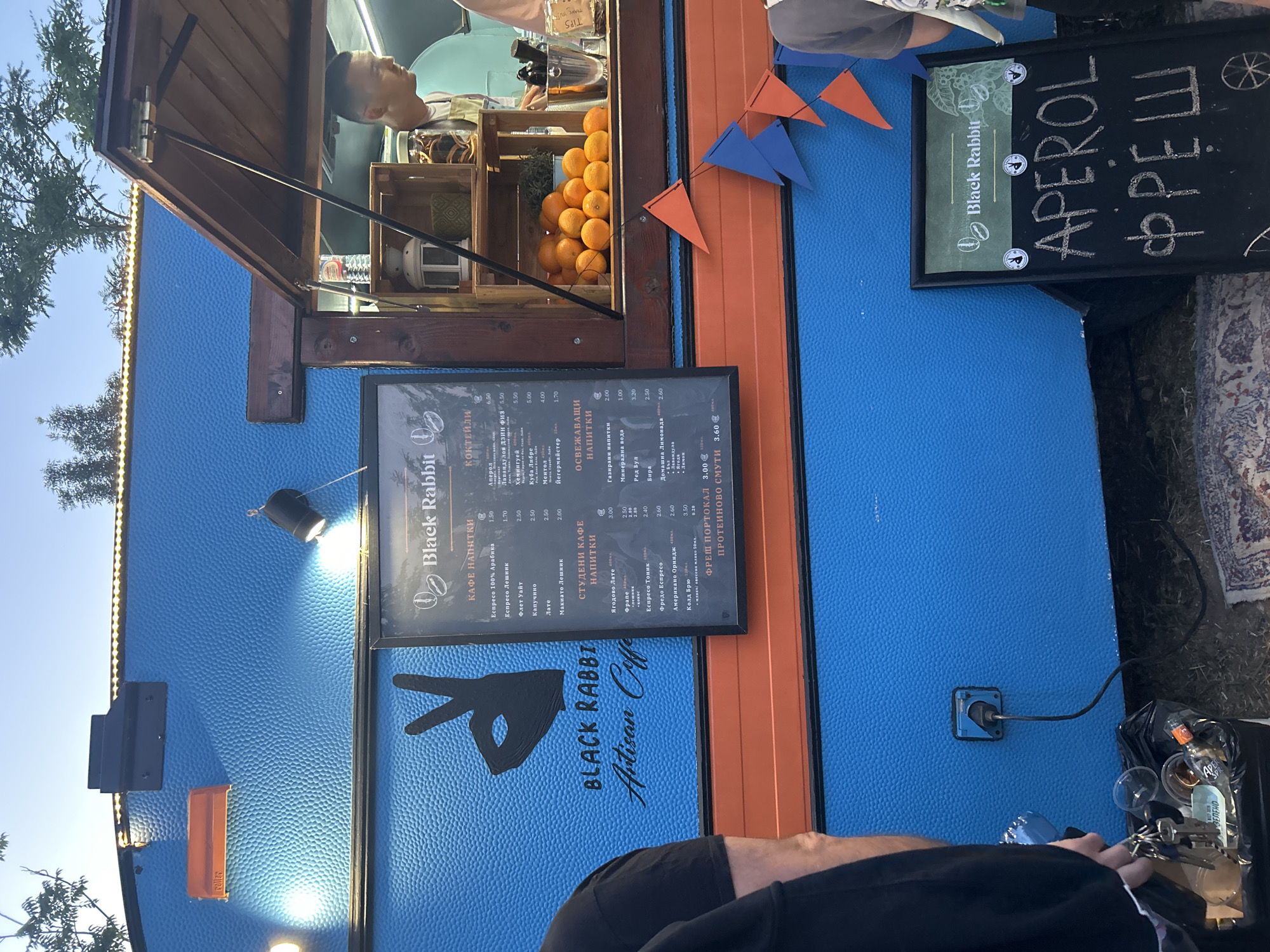 A brightly lit blue food truck with orange trim is parked outdoors, possibly at an event or market, with trees visible in the background under a clear sky. A black chalkboard sign in front of the truck advertises "APEROL ФРЕШ" with a logo above it. The food truck has a serving window where a man in an apron stands. Inside the window, oranges are visible in a wooden crate. A menu board is attached to the truck, listing various coffee drinks and cocktails. A power cord is plugged into an outlet on the side of the truck, leading to a trash can filled with disposable cups and other debris. Decorative orange and blue triangular flags are strung across the front of the truck.
