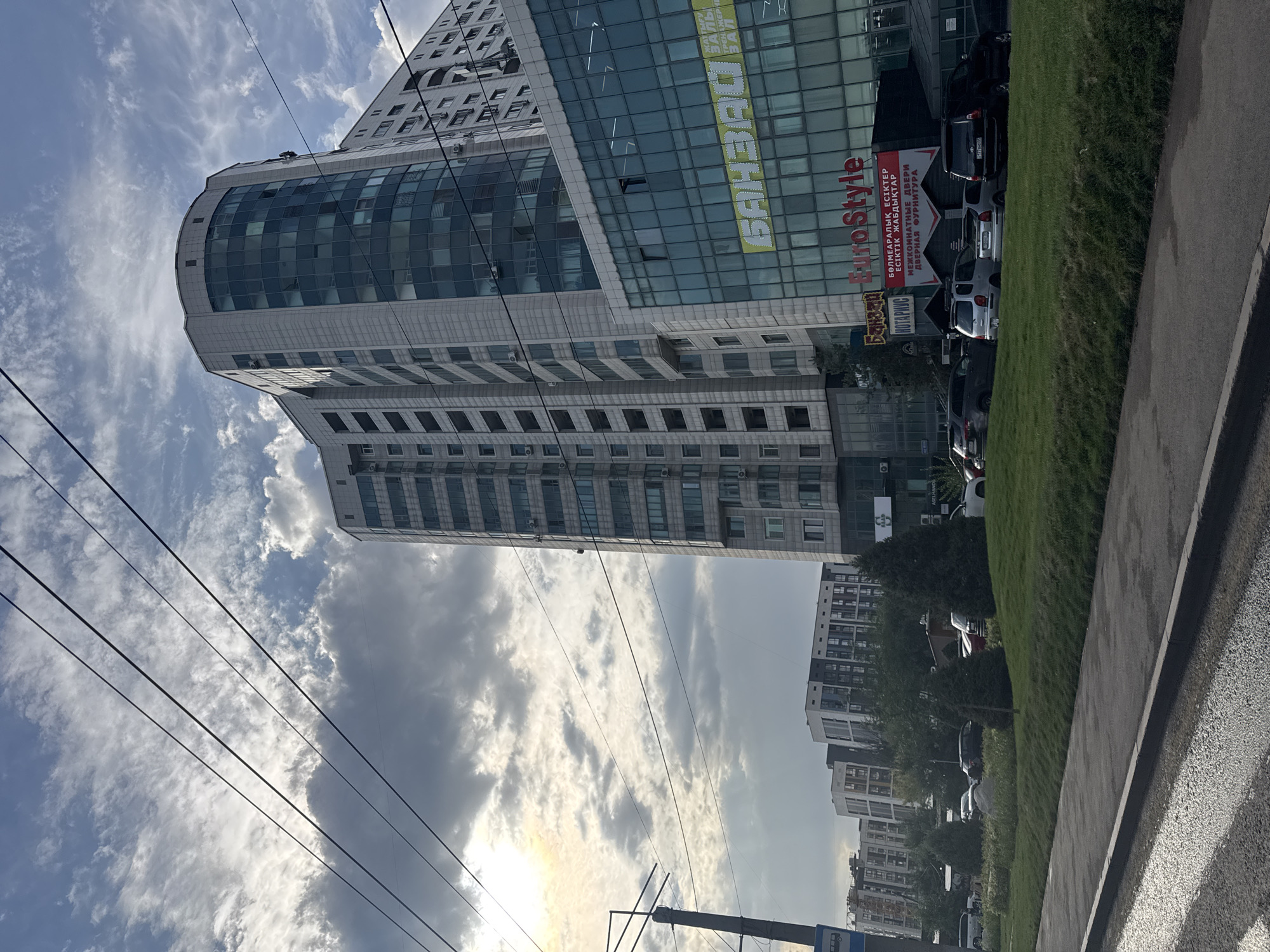 The image depicts a street scene in Almaty, Kazakhstan, with a focus on modern architecture and urban infrastructure under a cloudy sky. In the foreground, a paved road with white lane markings leads towards the right. A grassy median runs parallel to the road, dotted with evergreen trees.
Dominating the mid-ground and background are several multi-story buildings. The most prominent is a large, curved, modern building with a facade of light-colored panels and numerous blue-tinted windows. To its right, another building features a glass facade with visible signage. One sign, in yellow, reads "БАНЯ", and below it, in red, "EuroStyle". Text in red also indicates "INTERIOR DOORS". Further signs in red and white, in Cyrillic, appear to refer to services such as "FITNESS HALL" and "INTERIOR DOORS". Several cars are parked in front of these buildings.
To the left of the main buildings, a row of apartment buildings with a more modular design is visible, featuring white and dark gray facades and balconies.
The sky is partly cloudy, with sunlight filtering through breaks in the clouds, creating a somewhat dramatic lighting effect. Overhead, dark power lines crisscross the frame, adding to the urban atmosphere. A street lamp post with traffic signage, including what appears to be a bus stop indicator, is visible on the far left. The overall impression is of a busy, contemporary urban environment.