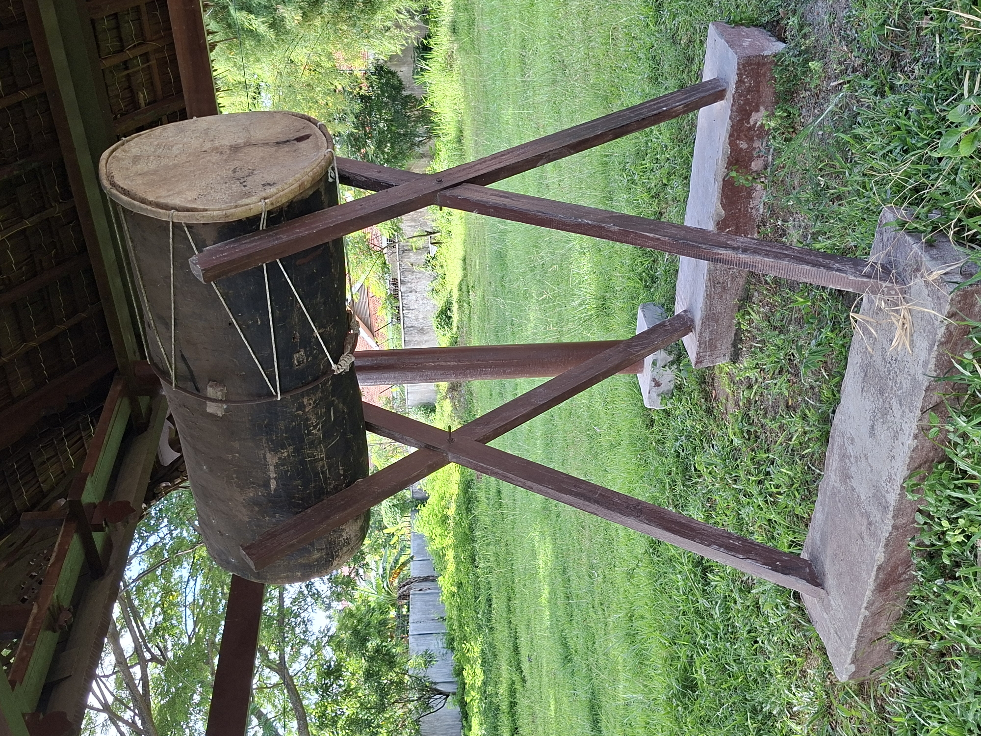 The image depicts a large, upright drum, likely a traditional Indonesian instrument, supported by a wooden stand. The drum has a cylindrical body, appearing dark in color, with a lighter-toned drumhead on top held in place by ropes. The wooden stand supporting the drum consists of two crossed wooden beams, held up by two rectangular stone structures. The setting appears to be outdoors, with a well-maintained lawn and various plants visible. In the background are trees and what seems to be a concrete wall with vegetation growing on it. Part of a building with a thatched roof is visible on the left side of the image. While the image is located in Lhokseumawe, Indonesia it does not have any people, actions, or interactions taking place.
