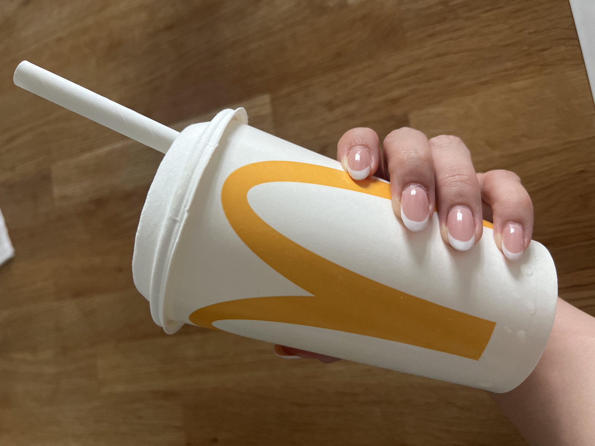 In Gdańsk, Poland, a close-up, high-angle shot shows a hand holding a white paper cup with the McDonald's logo. The cup contains a beverage and a white paper straw. The hand has a French manicure with clear polish. The background is a wooden surface, likely a table or floor. The lighting suggests an indoor setting, possibly a cafe or home.