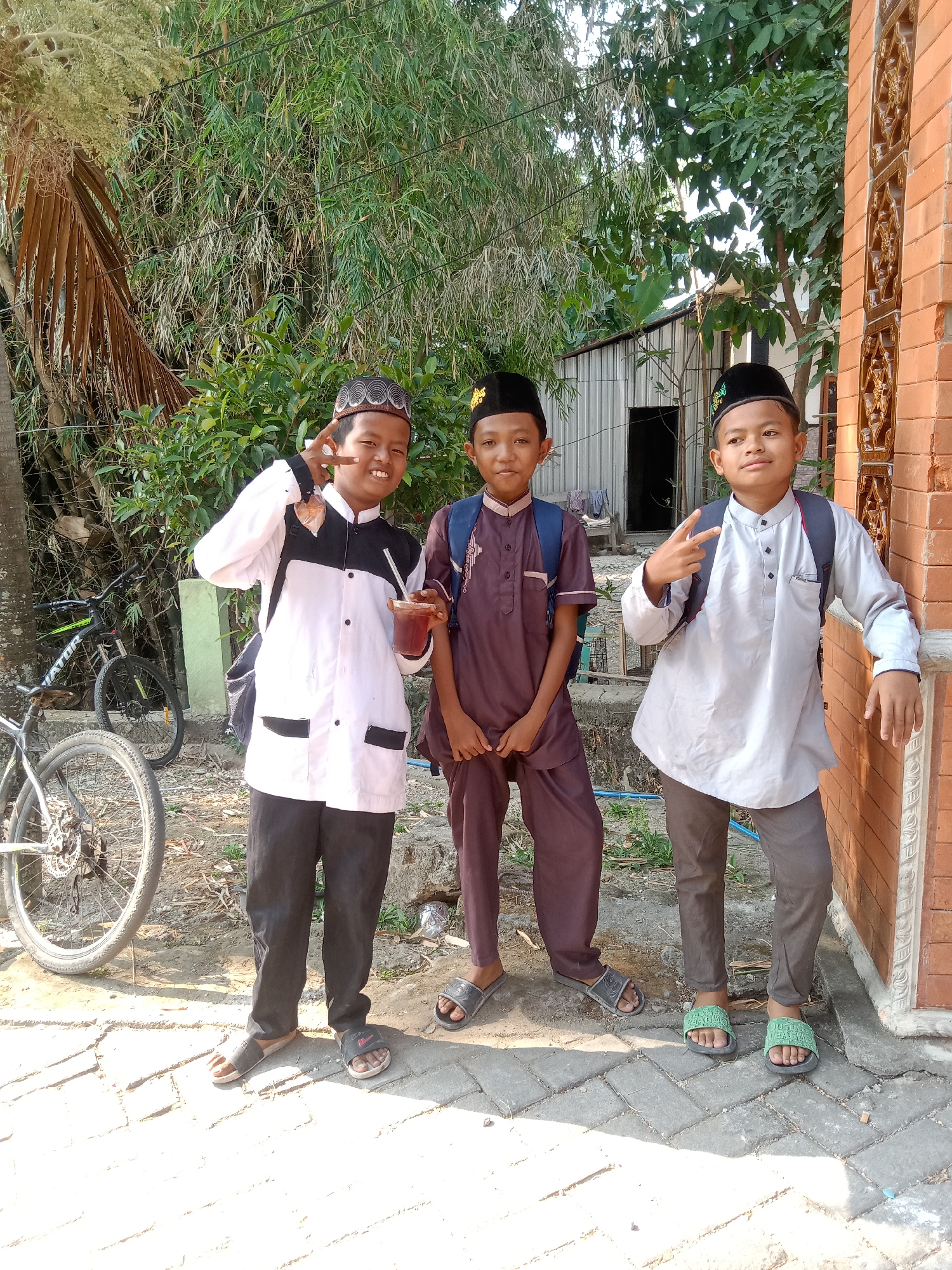Three young boys stand on a paved walkway in Stiaji, Indonesia. The boy on the left wears a white and black tunic, black pants, and a patterned kufi. He holds a drink with a straw and makes a peace sign with his left hand. The middle boy wears a dark brown tunic and pants, a black kufi, and a blue backpack. The boy on the right wears a light gray tunic, gray pants, a black kufi, and a dark backpack. He also makes a peace sign with his left hand. Behind the boys, there is dense green foliage, a palm tree, and a brick building. To the left of the boys, there are two bicycles. The scene is outdoors, with sunlight casting shadows on the ground.