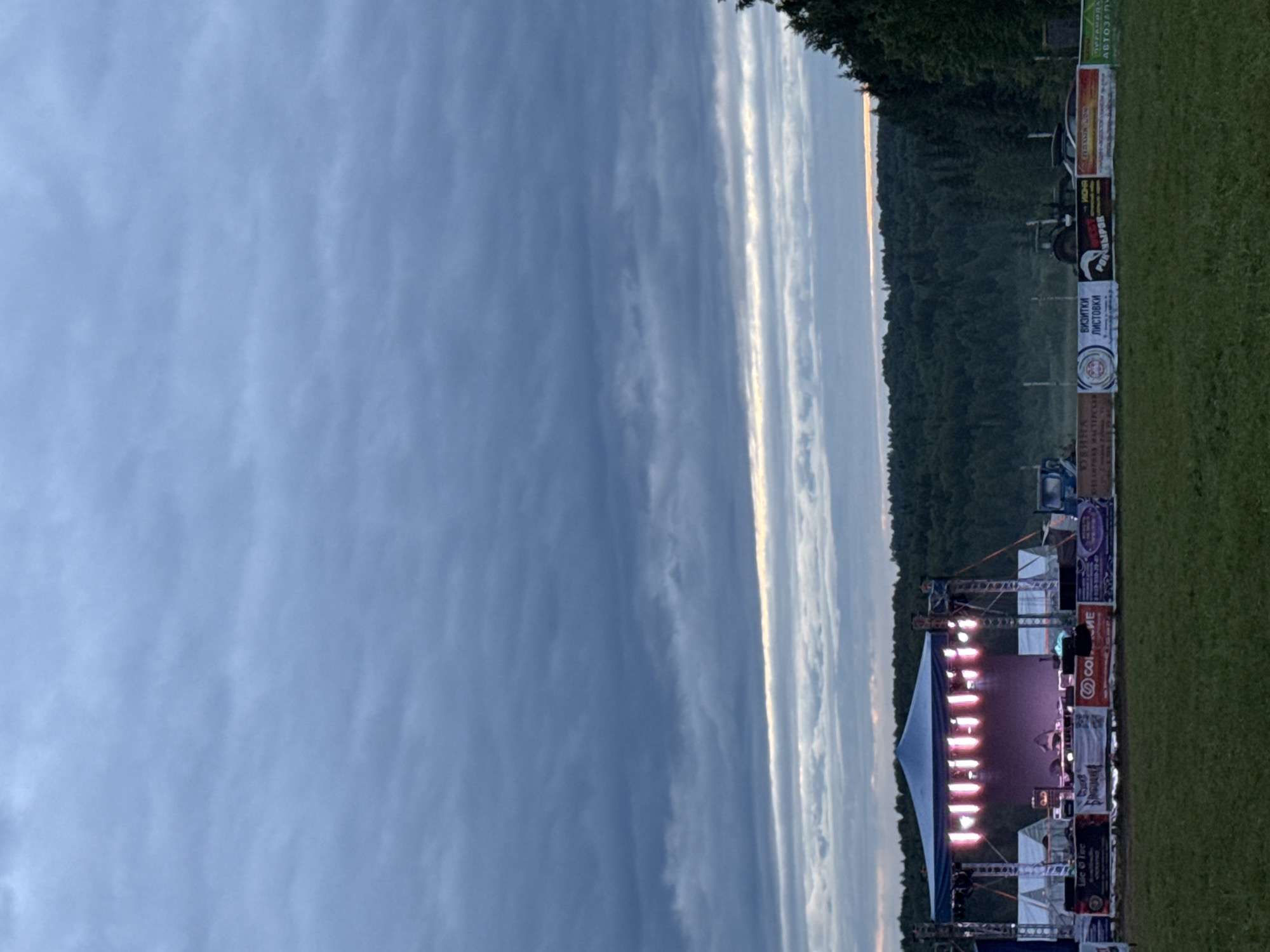 An outdoor event scene is depicted at dusk in Большая Кивара, Russia. The sky is largely overcast with dense, dark gray stratocumulus clouds dominating the upper two-thirds of the frame. A narrow, horizontal band of lighter, silvery-blue clouds with a subtle golden-orange glow is visible just above the distant tree line, indicating a fading sunset.
In the midground, a temporary outdoor stage is erected on a green grassy field. The stage structure consists of metal trussing supporting a dark blue canopy. A series of bright, pink-purple stage lights are active, illuminating the stage floor and backdrop. Visible on the stage are various pieces of sound equipment, including speakers and what appears to be a mixing console or control booth. Behind the stage, two white tents or temporary structures are set up.
Along the front edge of the stage area, several horizontal banners featuring advertisements are displayed. Some banners prominently display Cyrillic text, such as "БИЗИТКИ ЛИСТОВКИ" (Business cards, flyers). To the right of the stage and behind the banners, a dark green tractor and another vehicle, possibly a car or smaller truck, are parked in the grassy area. The background features a dense, dark green coniferous forest stretching across the horizon. The ground beneath the stage and in the immediate foreground is covered in green grass. There are no people clearly discernible in the image.