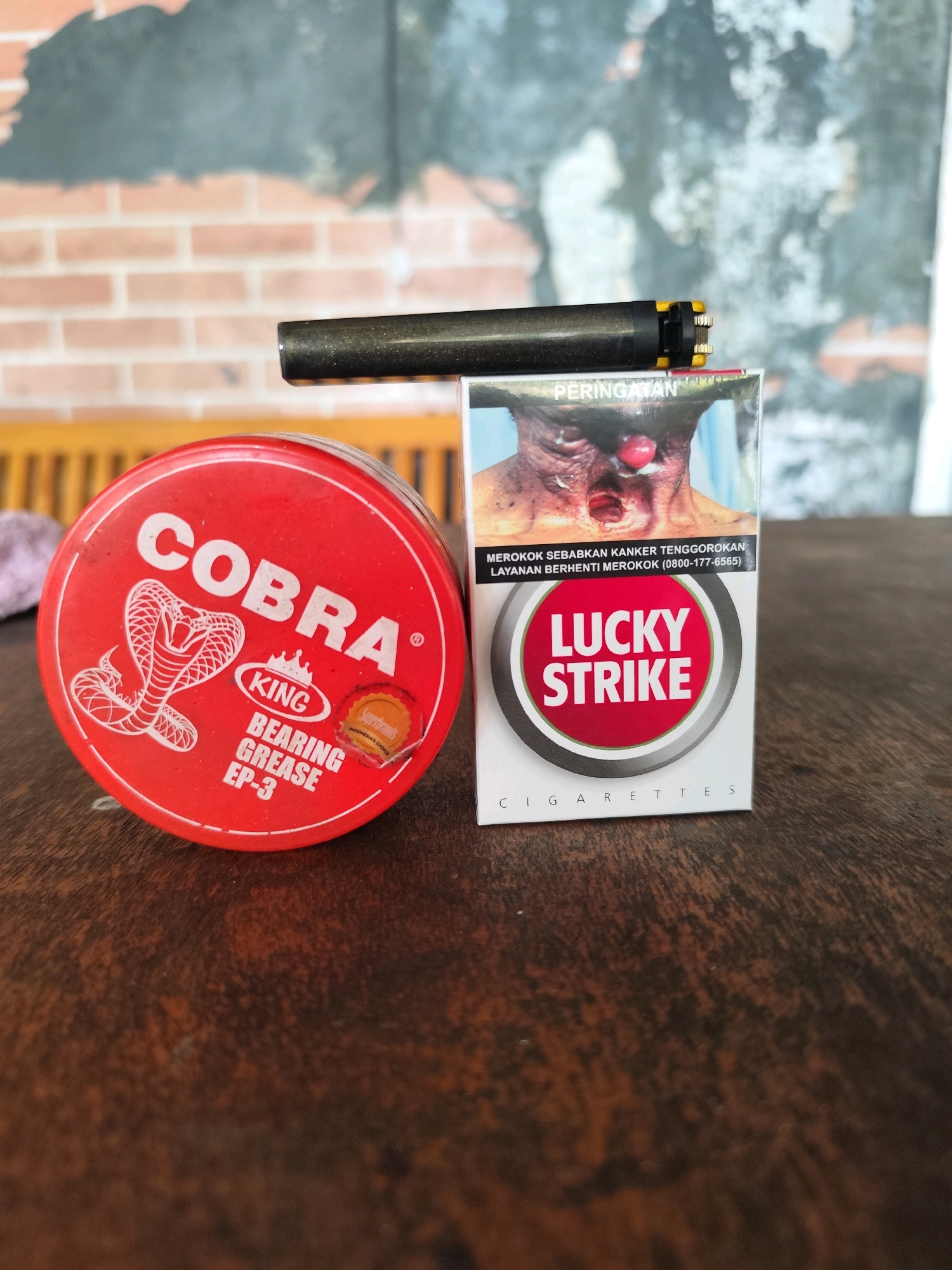 A Lucky Strike cigarette pack and a red tin of Cobra bearing grease are placed on a wooden table. A lighter lies across the top of the cigarette pack. The background shows a brick wall and a wooden bench. The scene is likely a close-up shot taken at an indoor or outdoor location in Palangka Raya, Indonesia, possibly on a table in a public eating area or a private home.