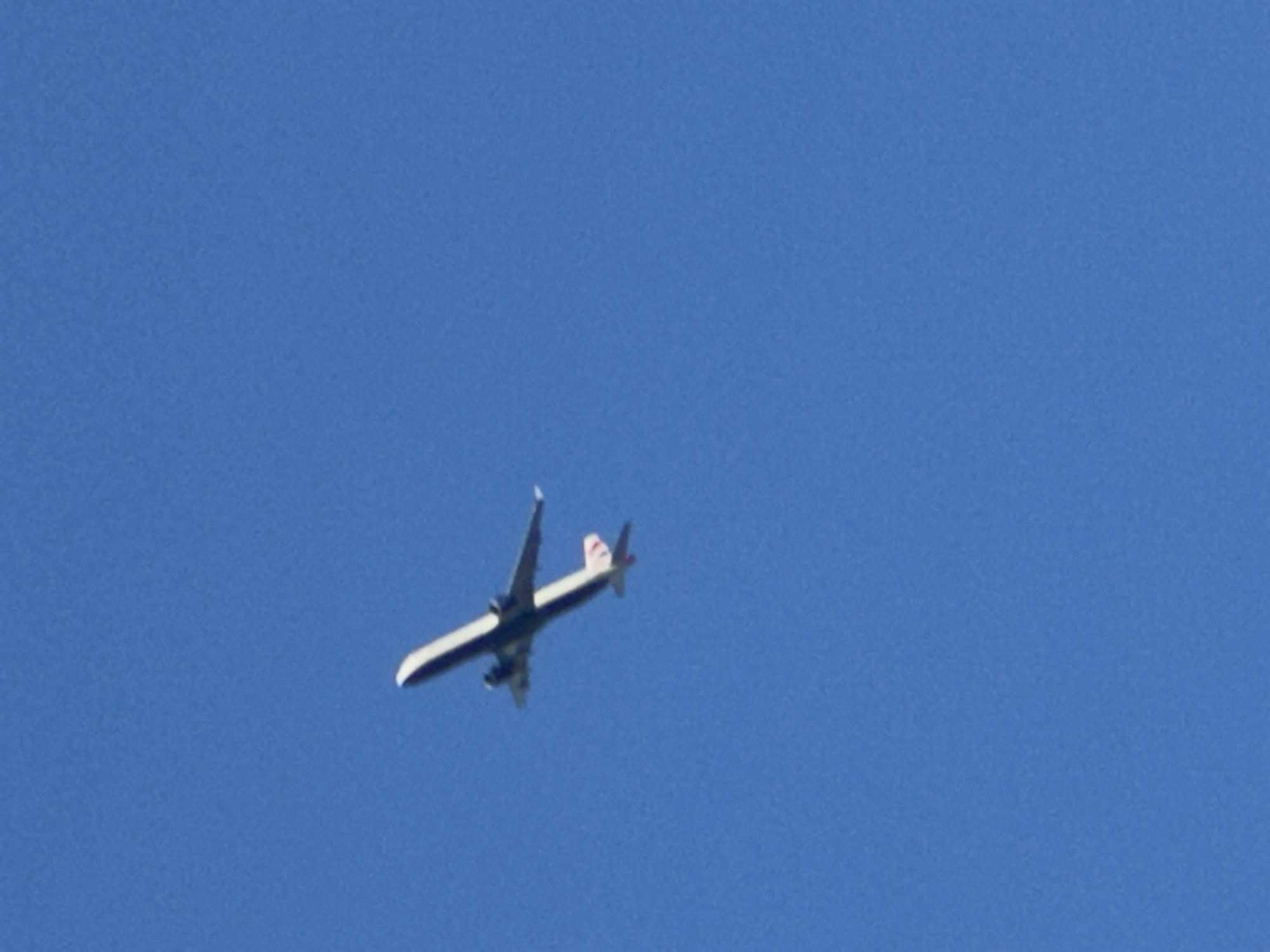 The image shows a passenger airplane flying in a clear blue sky. The airplane is seen from below and is angled slightly to the left. It is a commercial airliner with two engines mounted under its wings. The fuselage is white with a dark stripe along the side. The tail of the plane has a red and white emblem. The sky is a solid, uniform blue, indicating fair weather and likely daytime. There are no clouds visible. The image appears to be taken from the ground, looking up at the aircraft. The specific location of London, United Kingdom, is not discernible from the visual elements of the image itself. The time of day appears to be daytime, given the bright blue sky. There are no people, text, or specific location cues visible within the frame of the image. The activity taking place is simply the flight of an airplane.