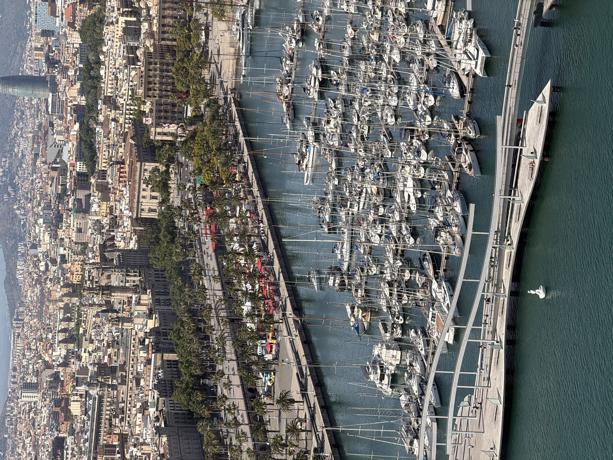 The image is an aerial view of the Port Vell marina in Barcelona, Spain. A large number of sailboats and yachts are moored in the harbor, with their masts forming a dense forest against the water. A modern wooden pier with curved white overhangs extends into the water, with several people walking and sitting on it. In the foreground, a small white sculpture of a person is visible in the water.
Behind the marina, a wide promenade lined with palm trees separates the water from a busy street with cars and some red-roofed cafes. Further inland, the cityscape of Barcelona unfolds, characterized by a mix of historic buildings with ornate architecture and more modern structures. Notable buildings include churches with spires and domes, and in the distance, the distinctive Torre Glòries skyscraper. The background shows a hilly, hazy landscape, typical of the Mediterranean. The sky is clear and blue, indicating a sunny day.