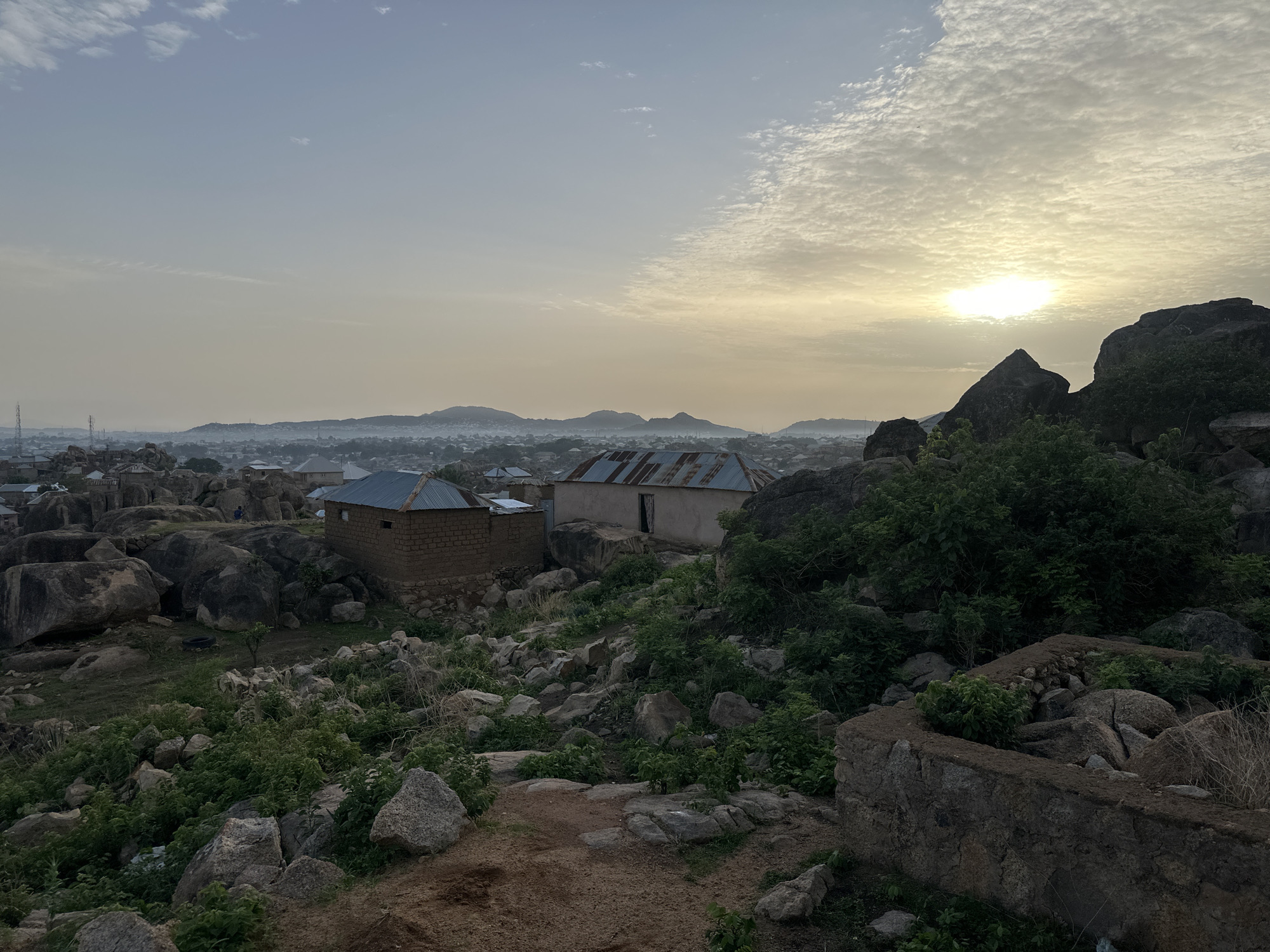 The image shows a rocky, elevated landscape with a scattering of buildings in the city of Jos, Nigeria. Two rudimentary structures with corrugated iron roofs are visible amidst the rocks and vegetation. The foreground consists of a path, rocks, and sparse greenery. In the distance, the city of Jos stretches out, with hills visible on the horizon under a cloudy sky where the sun is breaking through. The overall light suggests either early morning or late afternoon. No people are visible in the immediate scene. The general environment appears rugged and undeveloped.