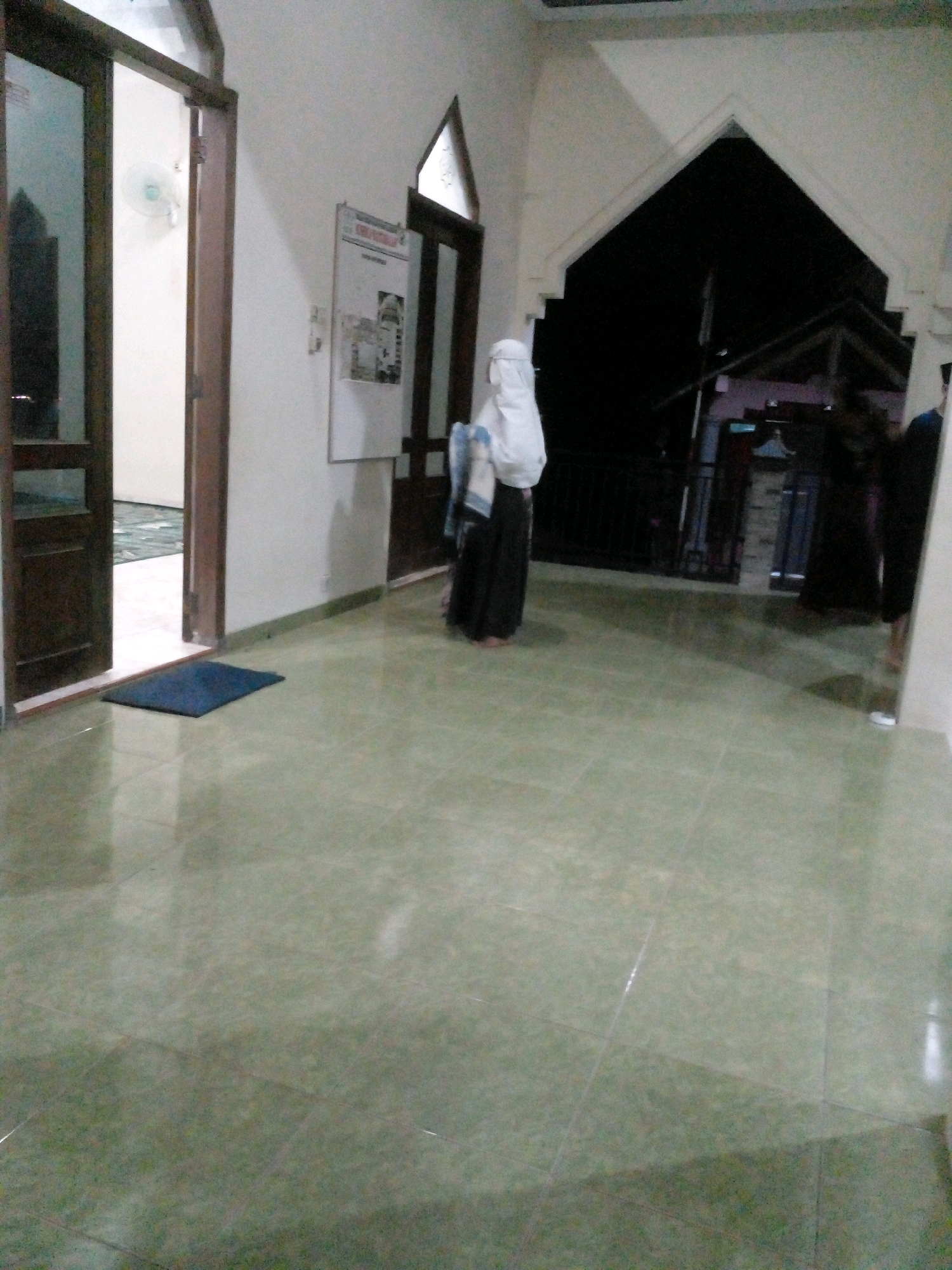 The image shows a hallway with light green tiled flooring. On the left, a dark wooden door stands ajar, revealing a room with a white wall and a ceiling fan. A dark blue mat rests on the floor in front of the door. Further down the hallway, a person in a white headscarf and black clothing stands with their back to the camera. To the right of this person, an arched opening leads to an outdoor area with a dark sky and silhouetted buildings. On the wall to the left of the arched opening, a bulletin board with papers is attached. The scene appears to be an interior space, possibly part of a mosque or religious building, in Pungangan, Indonesia.