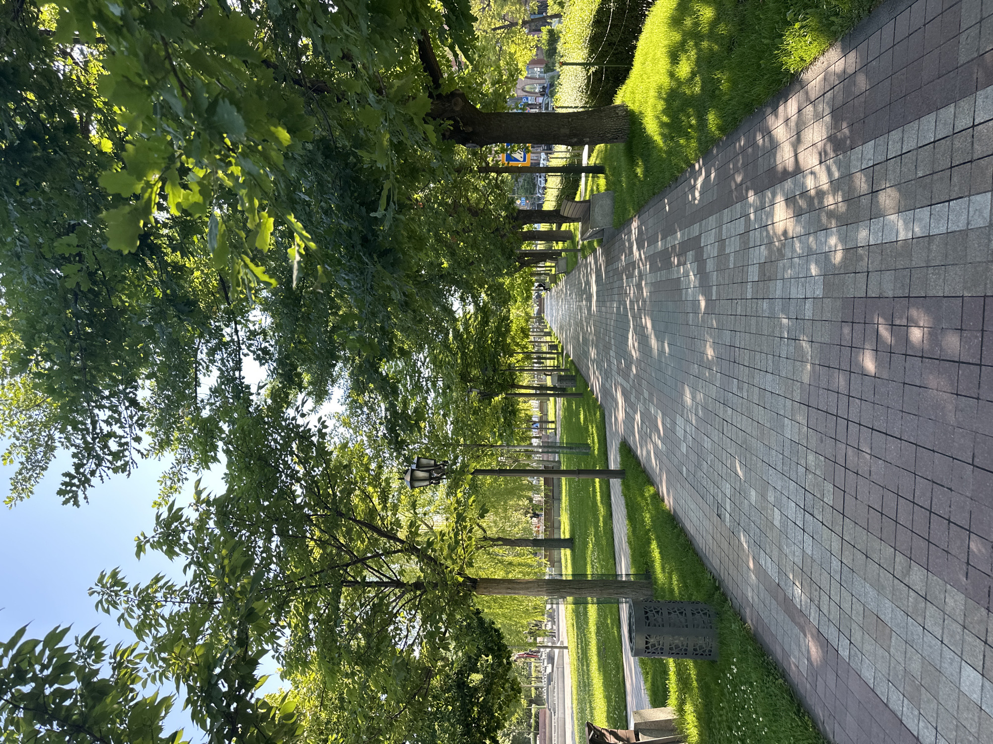The image depicts a tree-lined walkway in a park, likely in Yerevan, Armenia. The walkway is paved with light and dark grey/brown square tiles and stretches into the distance. Rows of trees with green foliage line both sides of the path. Street lamps are interspersed among the trees. Green grass borders the walkway. Benches are visible along the path, as well as trash bins. Shadows from the trees fall across the walkway. In the background, some buildings and a small structure can be seen. The sky is visible through the trees.