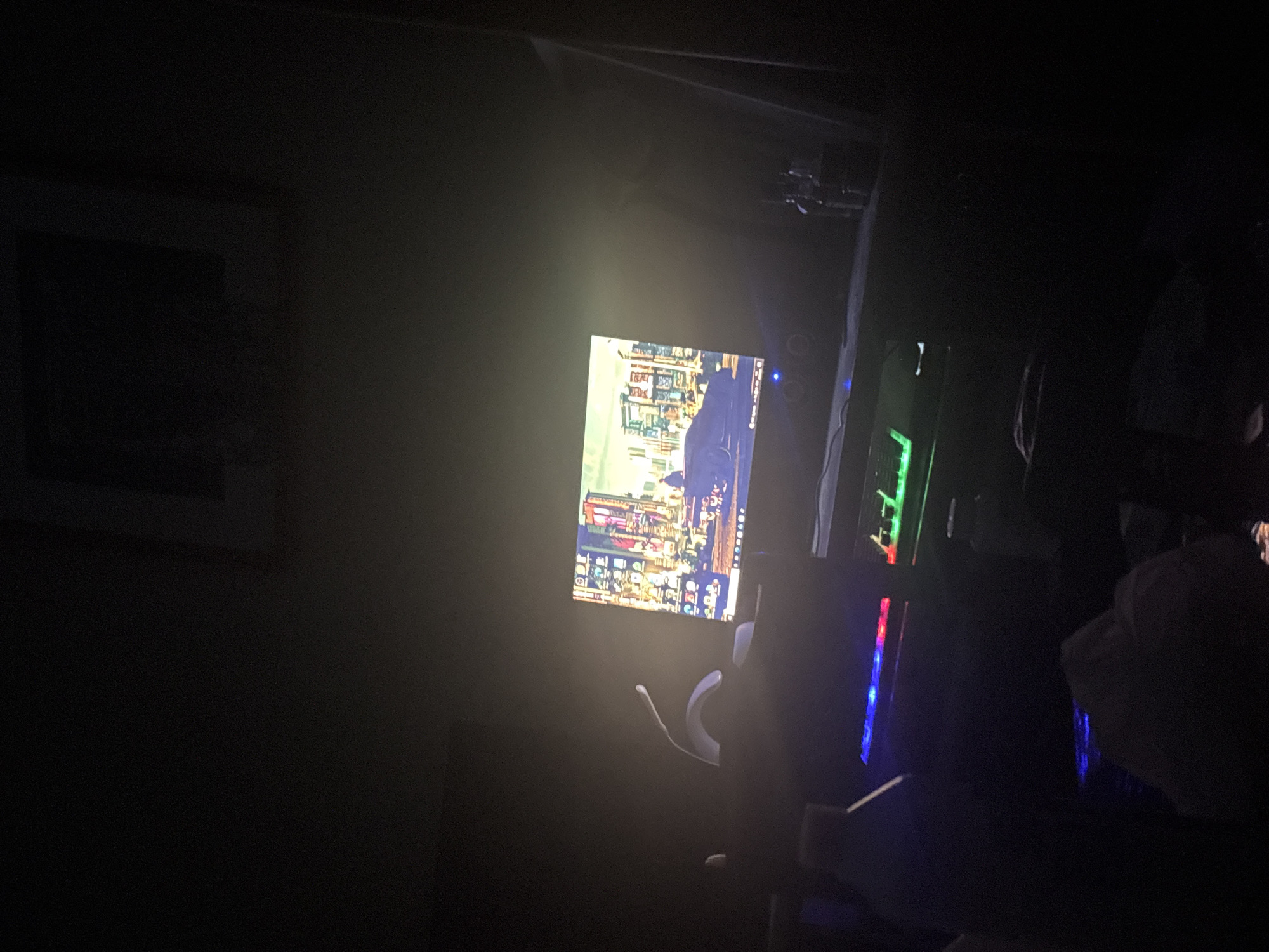 The image shows a dimly lit room with a computer setup. A computer monitor displays an image of a city, although the specific city is not identifiable from the visual information. Adjacent to the monitor is a computer tower with visible internal components and illuminated with RGB lighting. A headset is placed in front of the monitor. The overall environment is dark, suggesting a nighttime setting or a room with limited lighting. The image suggests an indoor setting with a focus on a gaming or computer workstation. The presence of a framed picture is noticed in the background.