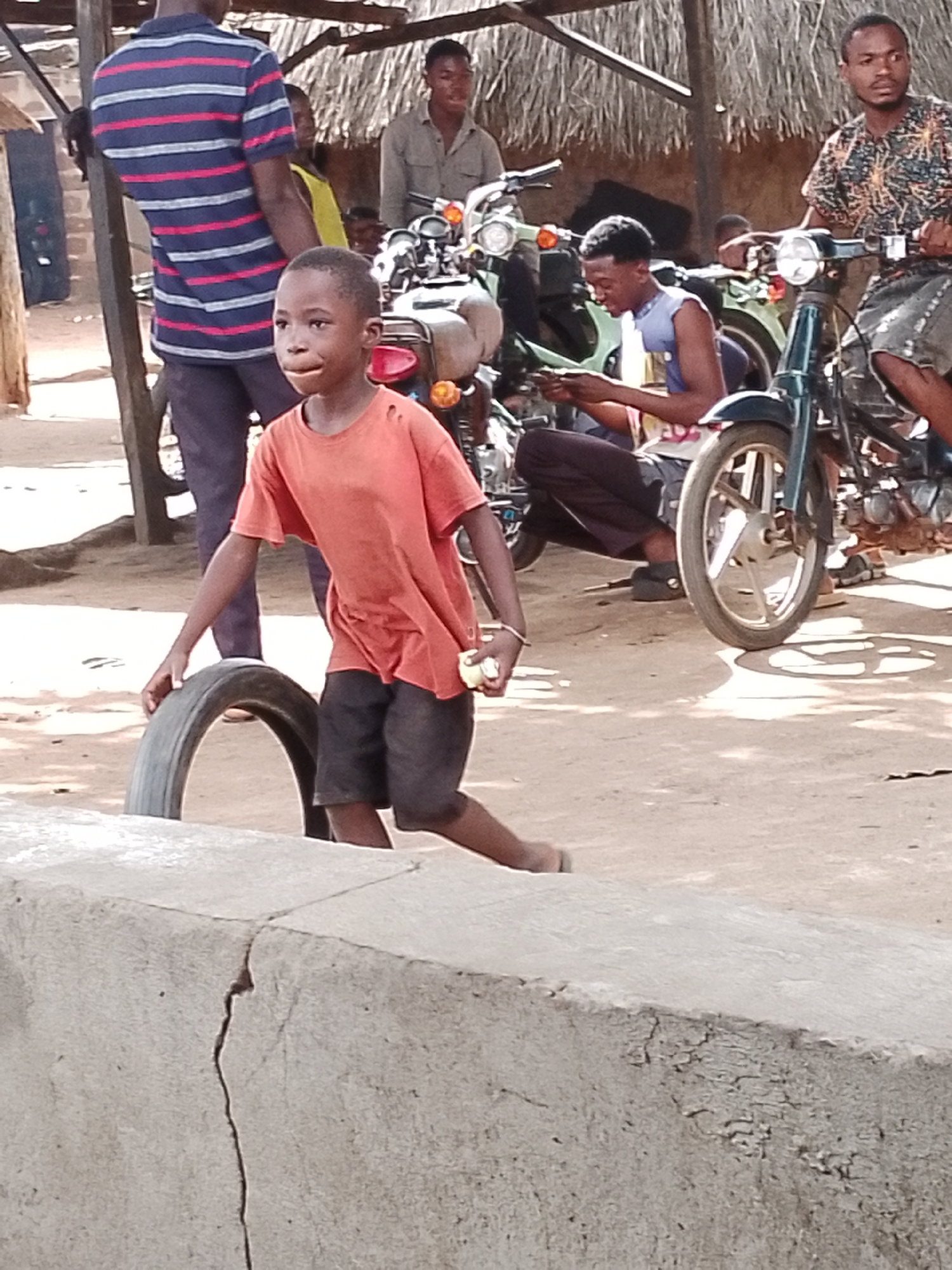 A young boy, wearing an orange t-shirt and dark shorts, runs with a tire in an outdoor, dirt-ground setting. He is on the right side of the frame, his body turned to the left and his head looking towards the left as well. His right hand is extended towards the tire, and his left arm is bent at the elbow. The tire is a dark, worn-out rubber with a visible tread pattern.
In the background, several people and motorcycles are visible. A man in a striped blue and red shirt stands to the left, his back to the camera. Behind him, another person in a yellow shirt is partially visible. Further back, two young men are seated on or near motorcycles. One is wearing a patterned shirt and appears to be on a motorcycle, while another is wearing a blue tank top and dark pants, looking down at something in his hands. Multiple motorcycles, some with visible handlebars and wheels, are parked in the area, suggesting a gathering or marketplace. The scene appears to be under a thatched roof structure, providing shade. The lighting suggests it is daytime. The ground is dusty and uneven. The overall atmosphere seems casual and active, possibly indicating a playful moment or a functional task involving the tire. There is a cracked concrete structure in the foreground that extends across the bottom of the image.