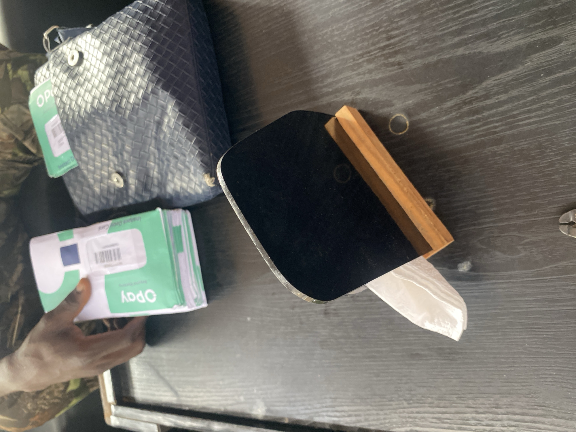 A hand is holding a stack of OPay branded debit cards, with "Instant Debit Card" printed on them. To the right of the cards is a dark woven bag, with a corner of another OPay card peeking out. In the foreground, a black rectangular object with a wooden base holds a white folded napkin. The scene appears to be set on a dark wooden table, possibly in a commercial area within Howayi, Nigeria, given the OPay branding, a mobile payment service popular in the country.