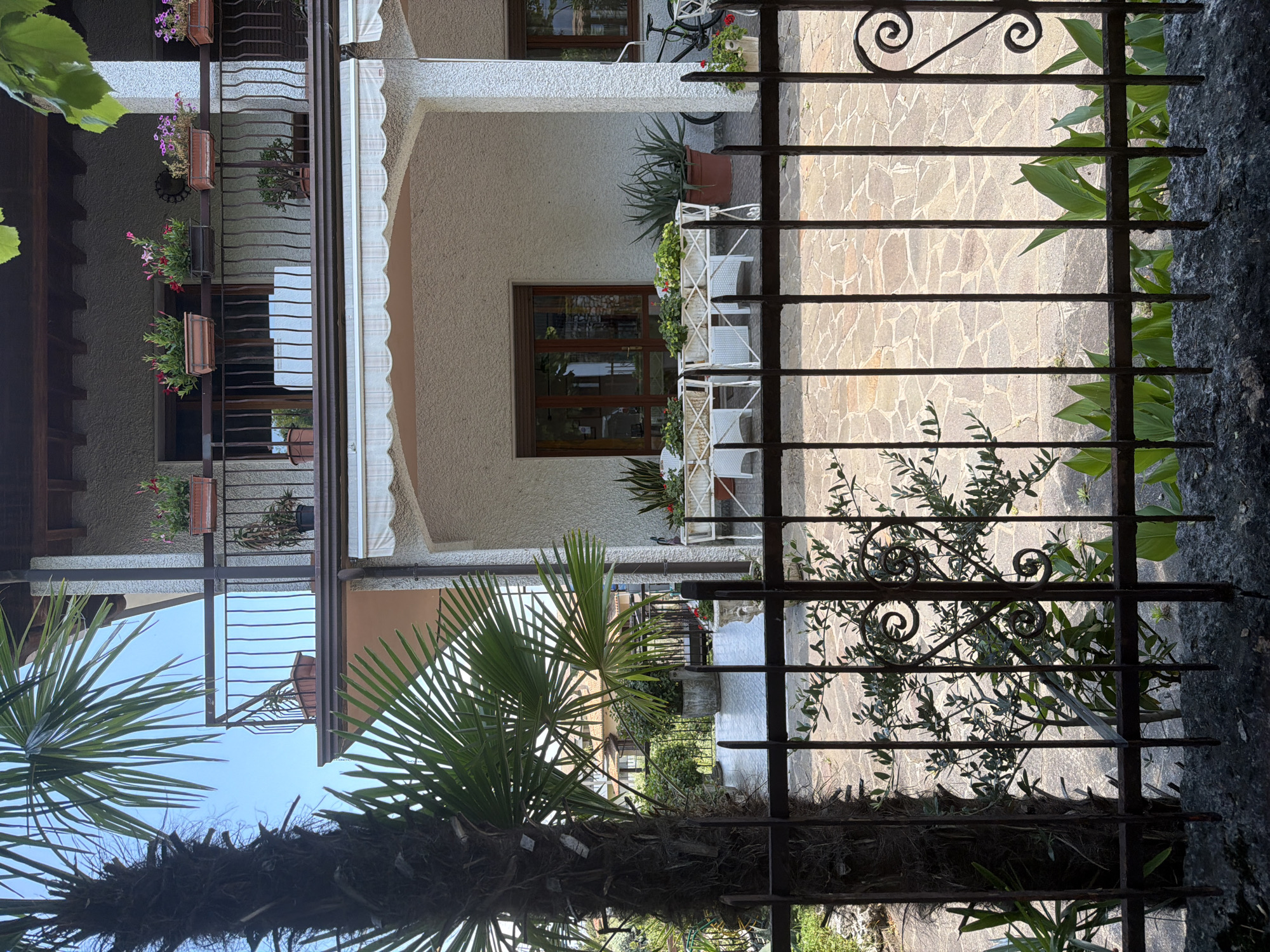 This image depicts the exterior of a multi-story building, likely a residence, in Padenghe sul Garda, Italy. The foreground features a dark metal fence with decorative scrollwork, partially obscuring a paved patio area. Beyond the fence, a large palm tree trunk is visible on the left, with its fronds reaching upwards and obscuring parts of the building.
The building itself is textured with a light-colored stucco finish. A balcony with a dark metal railing is visible on the upper level, adorned with several potted plants containing colorful flowers and greenery. A white awning extends from the lower level of the building, providing shade over a covered porch area. This porch features large glass doors and windows, with a table and chairs set up outside, also surrounded by potted plants.
To the right of the porch, a black bicycle is leaning against the wall, and more potted plants with vibrant red flowers are present. The ground on the patio is paved with irregular, flat stones in various shades of tan and grey. Behind the building, other residential structures with orange tiled roofs are visible, suggesting a neighborhood setting. The overall environment appears to be sunny and well-maintained, typical of a residential area in a Mediterranean climate.