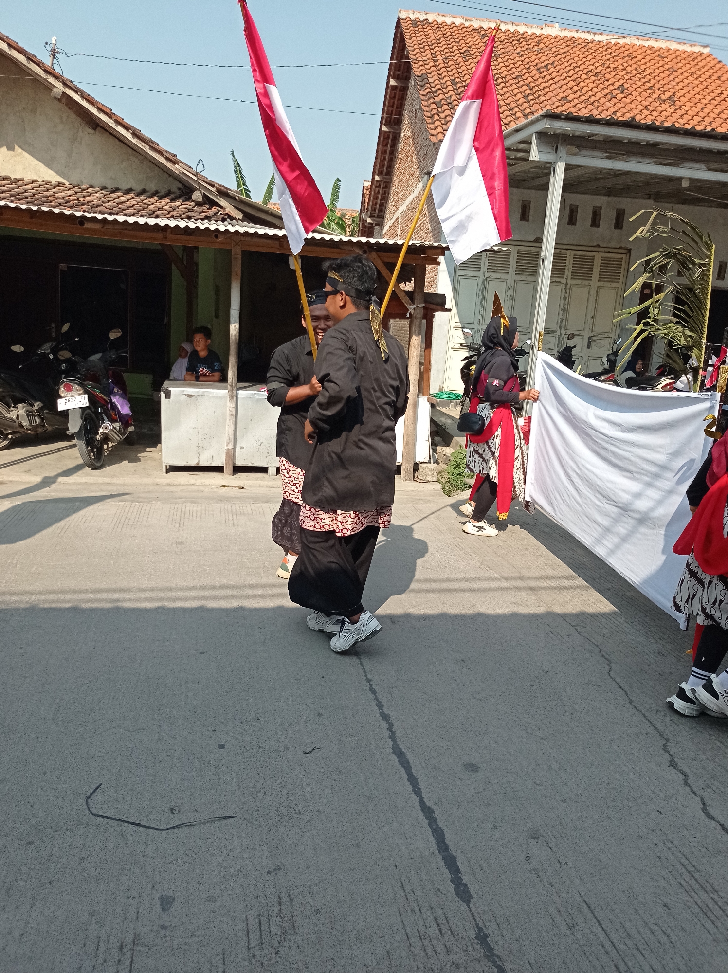 Two individuals in traditional attire are walking in a parade in Maribaya, Indonesia. The person in front is wearing a black shirt and pants with a red and white patterned sash, and they are holding a red and white flag. Behind them, another person in similar attire is also holding a flag. Other participants are visible in the background, including someone holding a large white cloth. The scene takes place on a paved street in front of houses with red tiled roofs. Motorcycles are parked near the houses, and a person is seated in a small stall. The sky is clear and blue.