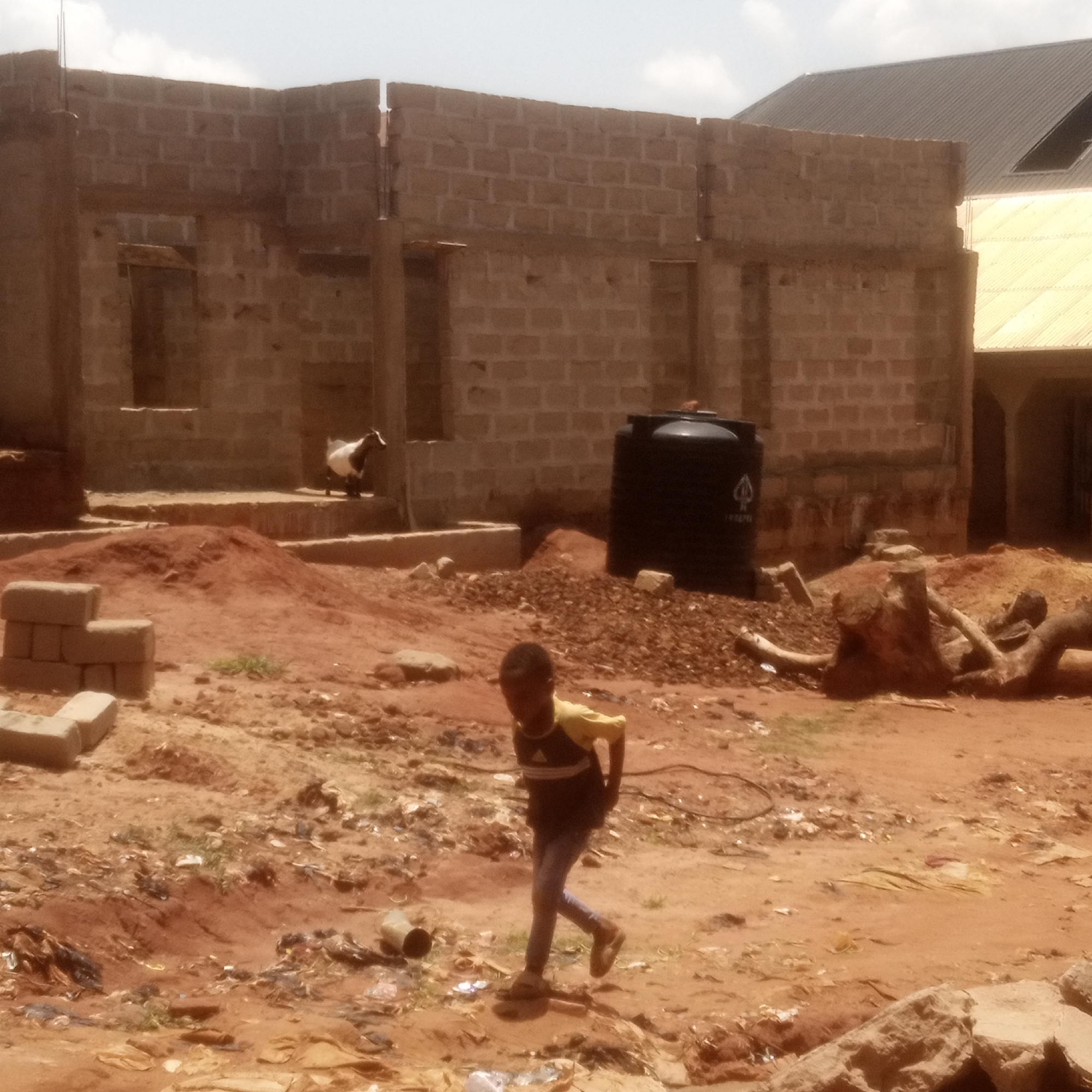 The image depicts a construction site in Unknown, Unknown. An unfinished brick building stands in the background. A black water tank sits near the building. A child is walking across the dirt and debris-covered ground in the foreground. A goat is perched on a ledge of the unfinished structure. Other objects include bricks, tree roots, and scattered trash.