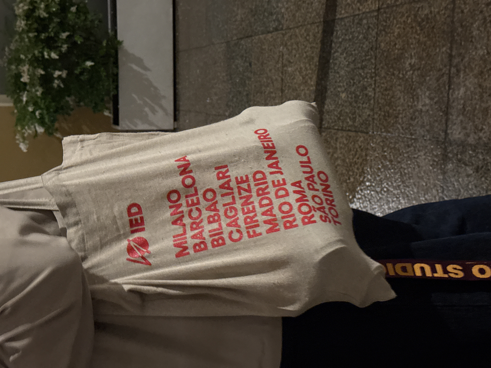A person is holding a tote bag in front of them. The bag is off-white with the word "IED" written in red, followed by a list of cities, also in red: MILANO, BARCELONA, BILBAO, CAGLIARI, FIRENZE, MADRID, RIO DE JANEIRO, ROMA, SAO PAULO, TORINO. The person is wearing dark blue pants and a belt. The surrounding environment is not clearly visible.