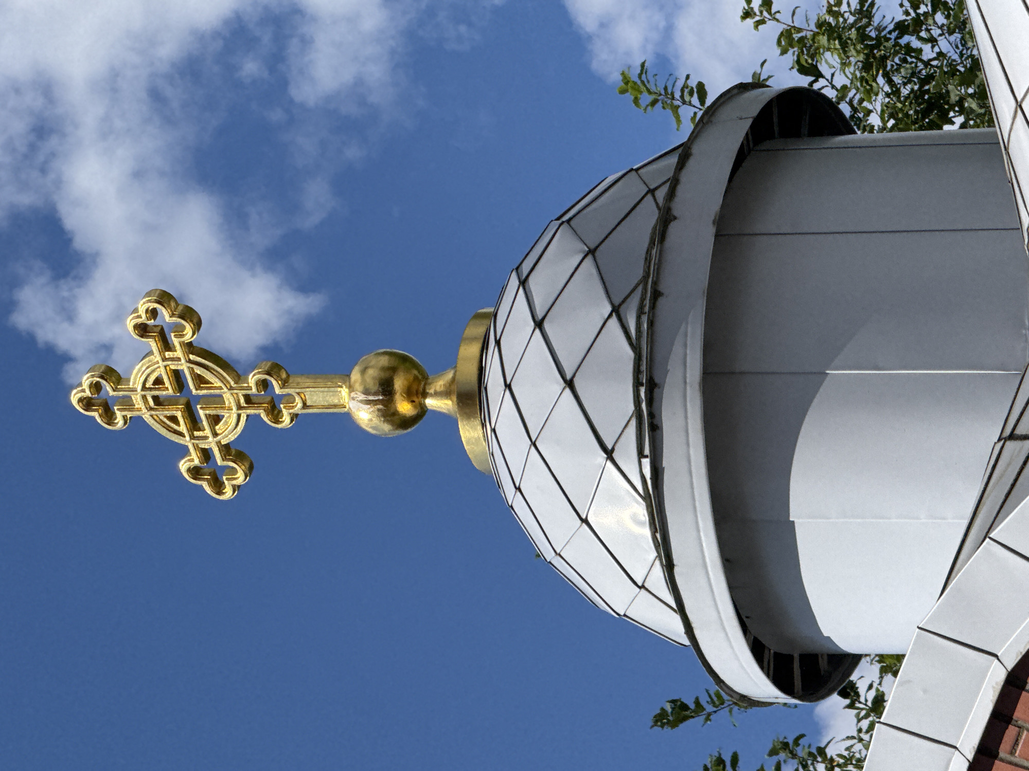 A close-up, low-angle shot captures the golden cross atop a domed structure, likely a church or chapel, against a bright blue sky with scattered white clouds. The cross is ornate and gilded, with decorative loops at the end of each arm. Below the cross, a golden orb sits on a golden base, which in turn rests on a light grey, diamond-patterned dome. The dome appears to be made of metal panels. The lower portion of the structure is a cylindrical, light grey section, with visible panel seams. Green foliage from nearby trees is visible at the edges of the frame, partially obscuring the dome. The overall impression is one of religious architecture, possibly in Saint Petersburg, Russia, given the context of the request for its location.