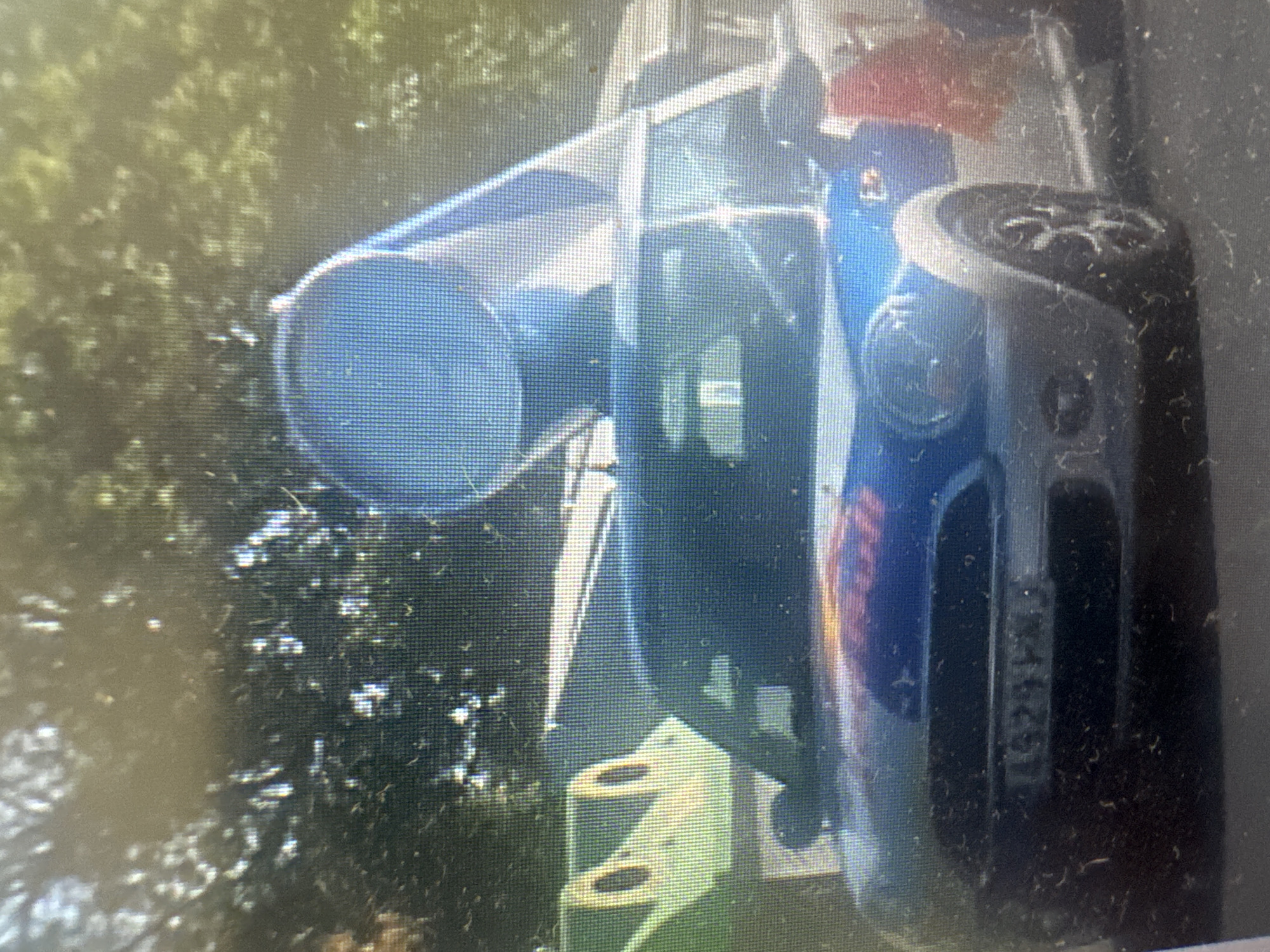 The image shows a blue and black object, possibly a car or vehicle, in an outdoor setting with greenery in the background. The object appears to be partially overturned or in an unusual position. There are reflections on the surface of the object, suggesting it is made of a smooth material. The lighting indicates it might be daytime, with some shadows visible. The overall scene is blurry and pixelated, making it difficult to discern fine details. Some of the provided crops highlight specific areas, like the wheel of the object, a window area with reflections, and a dark panel with what appears to be a license plate or identification number. The text "L6295" is visible on the dark panel. The setting appears to be a natural environment, perhaps a roadside or a wooded area.