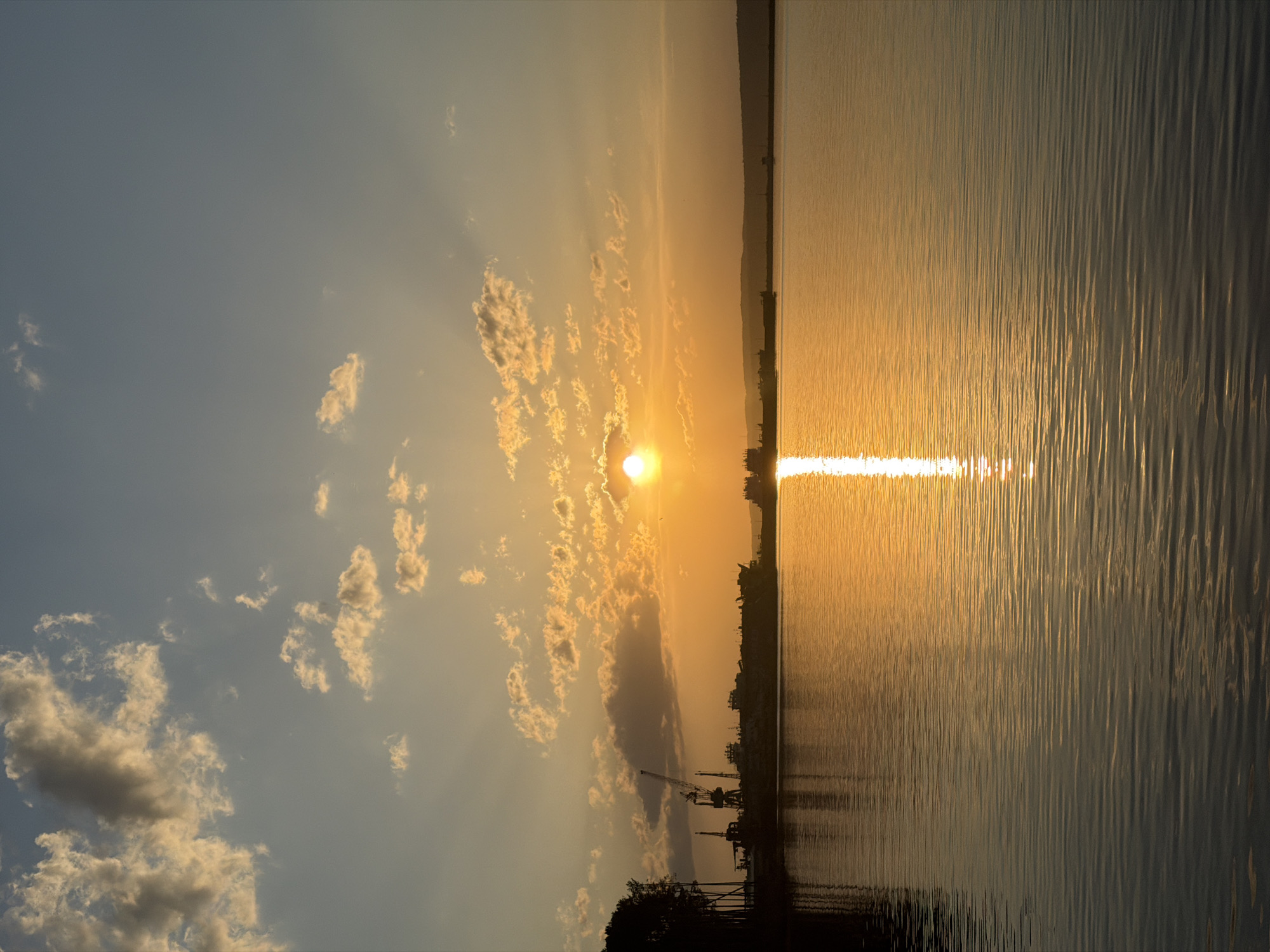The image depicts a sunset scene over a body of water, likely the Kama River, in Chaikovsky, Russia. The sun is partially obscured by clouds, creating rays of light. The reflection of the sun stretches across the water. A shoreline with vegetation and industrial structures, including cranes, is visible in the foreground. The sky transitions from light blue to orange-yellow near the horizon.