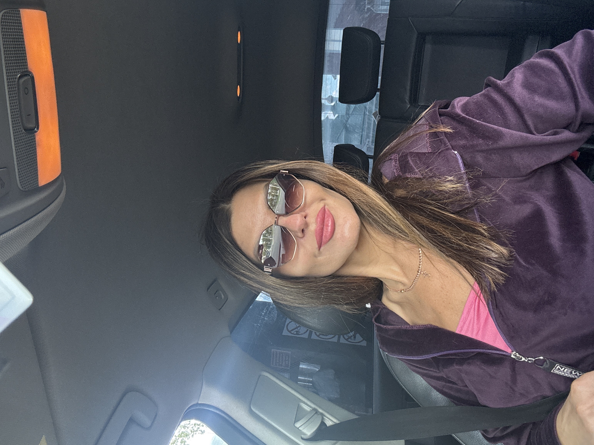 A woman is seated in the front passenger seat of a vehicle, taking a selfie or being photographed, with the location identified as the city of Chaikovsky, Russia. She has long, brown hair with lighter highlights and is wearing large, octagonal-shaped sunglasses with reflective lenses. Her lips are adorned with pink lipstick. She is wearing a dark purple velour hooded jacket, partially unzipped to reveal a bright pink top. A gold-colored chain necklace with a small cross pendant is visible around her neck. A black lanyard with "NEW" text is attached to her jacket zipper. A seatbelt is fastened across her right shoulder and chest. The car interior features a gray fabric headliner, with an orange-lit rectangular overhead light panel towards the front and two smaller orange-lit round lights towards the rear. Black leather or pleather seats and two rear headrests are visible. Reflections in the car's side window on the left clearly show the backward Russian text "ЗАПРЕЩАЕТСЯ КУРИТЬ" (Smoking is prohibited) along with "no smoking," "no food or drink," and "no littering" symbols, consistent with signage often found in public transport or taxis in Russia. The rear window reflection shows distant buildings, including structures with domes, indicative of a city environment.
