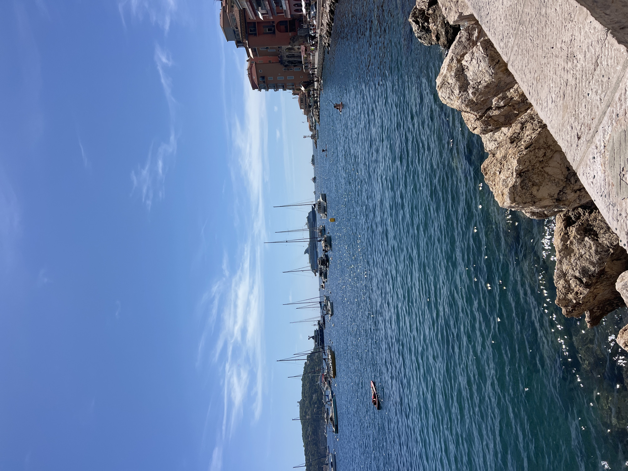 The image captures a sunny day at the port of Villefranche-sur-Mer, France, with the Mediterranean Sea occupying the foreground and mid-ground. The water is a deep blue with ripples reflecting the sunlight. In the foreground, large rocks are piled along a stone walkway. A person is visible lying on a red inflatable raft in the middle of the water. Further out, numerous sailboats and a large cruise ship are anchored. Along the right side of the image, colorful buildings with orange and red facades line the waterfront promenade, where people can be seen walking and sitting at outdoor cafes. The sky above is a clear blue with scattered wispy white clouds. In the distance, a green, tree-covered hill rises, topped with structures.