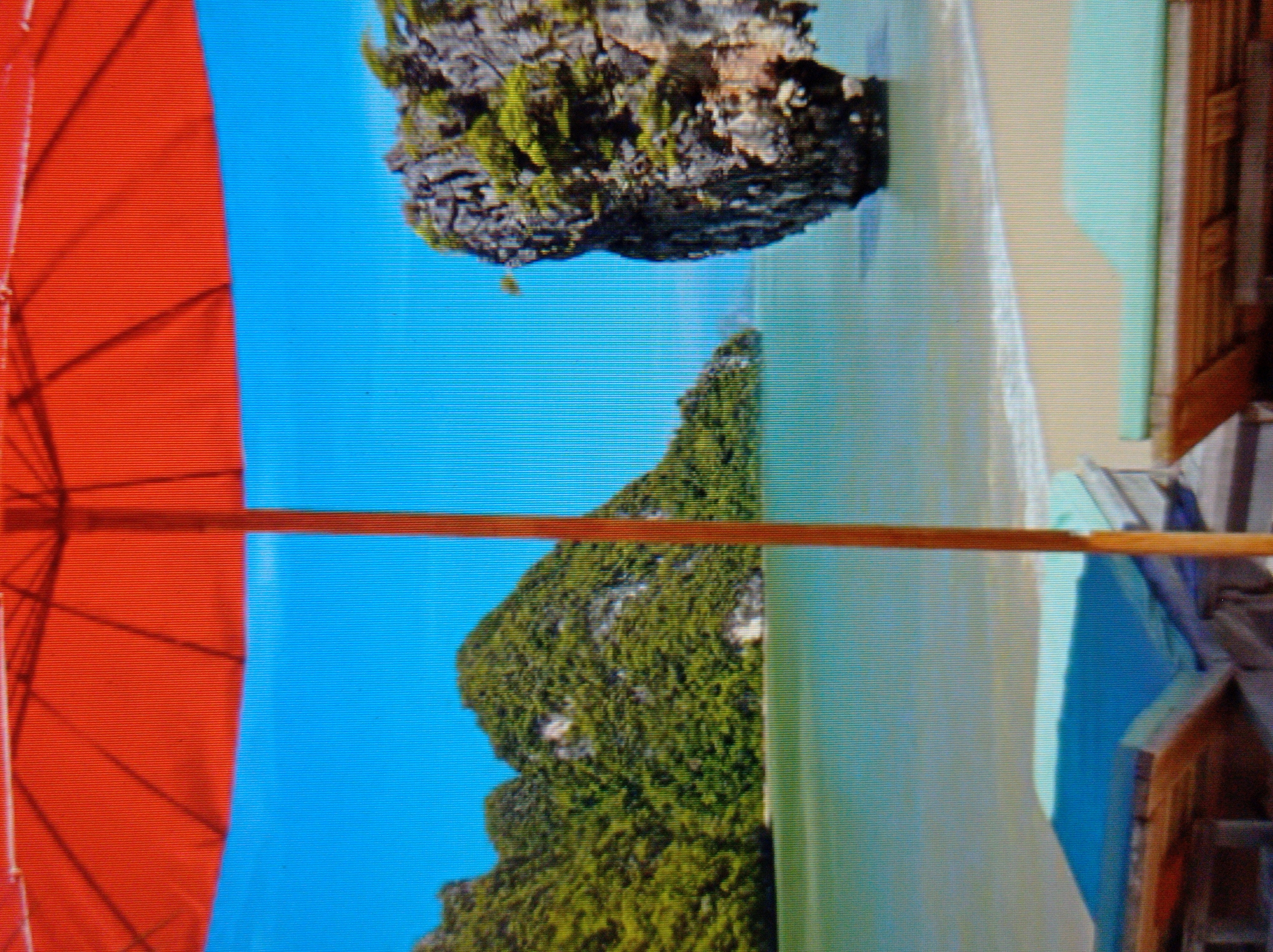 This image depicts a tropical beach scene, likely in Southeast Asia, featuring a large red beach umbrella in the foreground casting a shadow. The umbrella's pole is centrally located and extends downwards. Beyond the umbrella, a calm, turquoise body of water meets a sandy beach. Lush green, tree-covered islands or rock formations rise dramatically from the sea in the mid-ground and background. The sky is a clear, bright blue. In the lower foreground, there are parts of two wooden lounge chairs with light blue cushions, positioned on the sand. The scene suggests a relaxed vacation or resort setting. There is no indication of the city of Mukachevo, Ukraine, in this image; the environment is clearly not characteristic of that region.