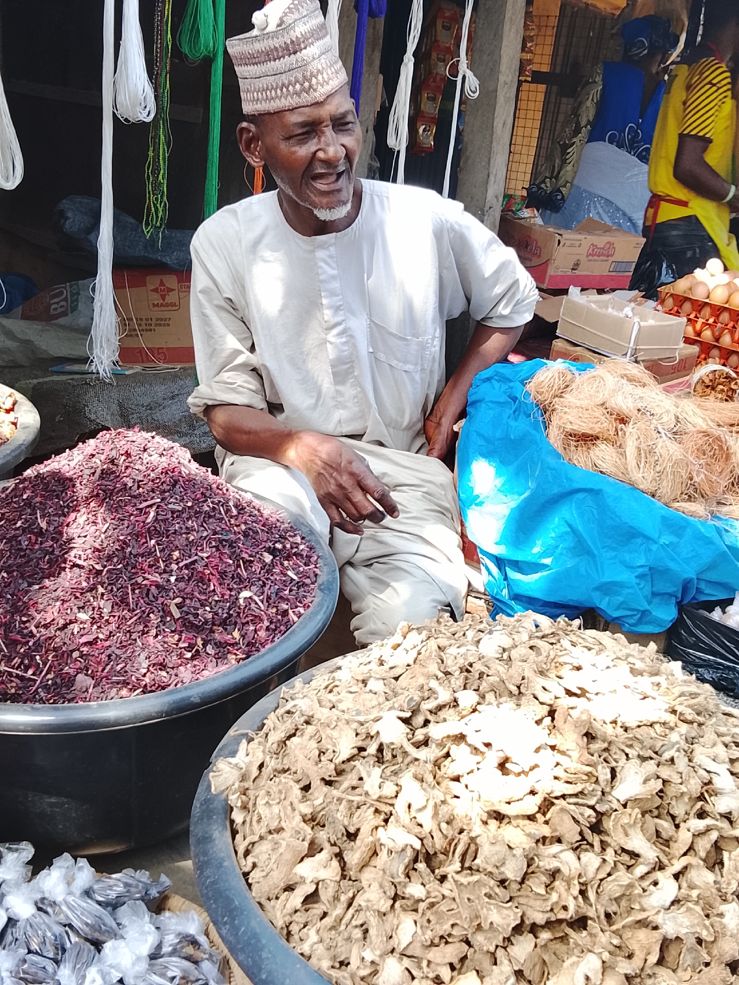 The image depicts an outdoor market stall in Akwanga, Nigeria. An older man, presumably a vendor, is centrally seated behind various goods. He wears a light-colored traditional long-sleeved top, light trousers, and a patterned white and brown cap. He has a white beard and mustache, and his mouth is open as if speaking or laughing. His right hand rests on his leg.
In the foreground, two large black basins contain dry goods. The left basin holds a substantial mound of shredded, reddish-purple material, consistent with dried hibiscus flowers. The right basin contains a large mound of dried, light brown, irregularly shaped pieces, consistent with dried ginger. Small, clear plastic bags filled with dark items are partially visible to the immediate left of the hibiscus.
Between the seated man and the dried ginger, a bright blue plastic bag holds numerous bundles of thin, light brown, fibrous material. In the background, behind the man and to his right, stacked cardboard boxes are visible, including one labeled "MAGCL" and another "Krasch." Below these boxes, multiple brown cartons of eggs are present. To the far right, a partially visible individual in a yellow and black striped shirt and a red apron stands near the eggs. Behind this individual, another person wearing a blue head covering and patterned blue garment is partially visible.
The market stall features vertical wooden support beams, and various white and green strands are suspended from above. The scene is illuminated by natural daylight.