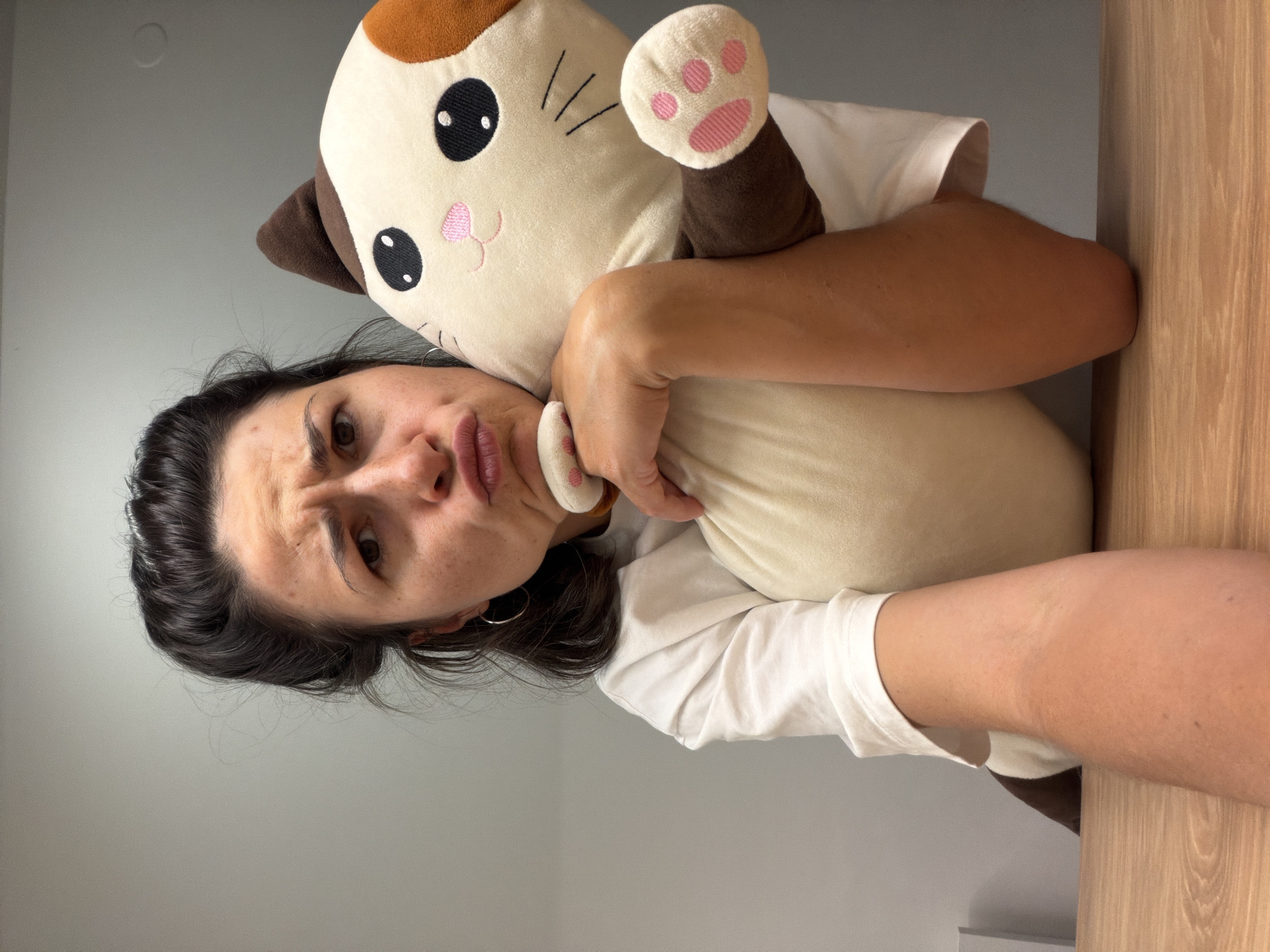 A woman with dark hair and light skin is holding a plush cat toy. She is wearing a white shirt and has a silver hoop earring in her left ear. Her facial expression is one of displeasure or mild irritation. The background consists of a light gray wall and a wooden surface on the right side of the frame. The location cannot be determined.
