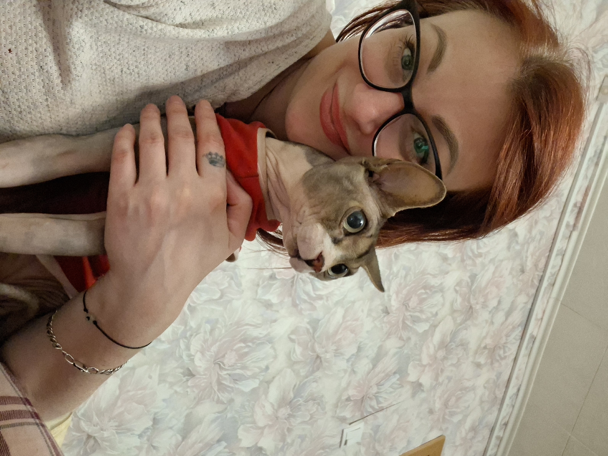 A close-up, selfie-style shot shows a woman with reddish-brown hair and green eyes, wearing black-rimmed glasses and a light-colored sweater, holding a hairless cat. The cat is grey and white with large, wide eyes and is wearing a red shirt. The woman is smiling, and the cat is looking off to the side. The background features floral patterned wallpaper. The scene appears to be indoors, likely in a residential setting, possibly in Забалоцкі сельскі Савет, Belarus.